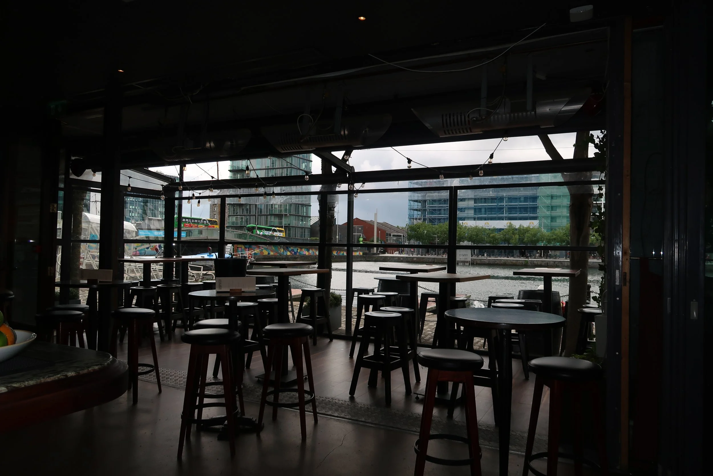 Interior of a restaurant with round tables and stools. Large windows reveal an outdoor scene with boats on water and modern buildings in the background.