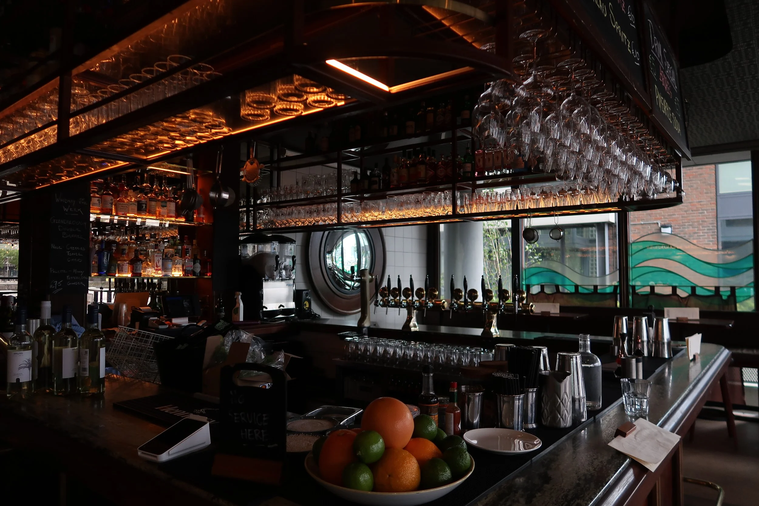 Interior of a bar with hanging glassware, bottles of alcohol, and a bowl of citrus fruits on the counter.