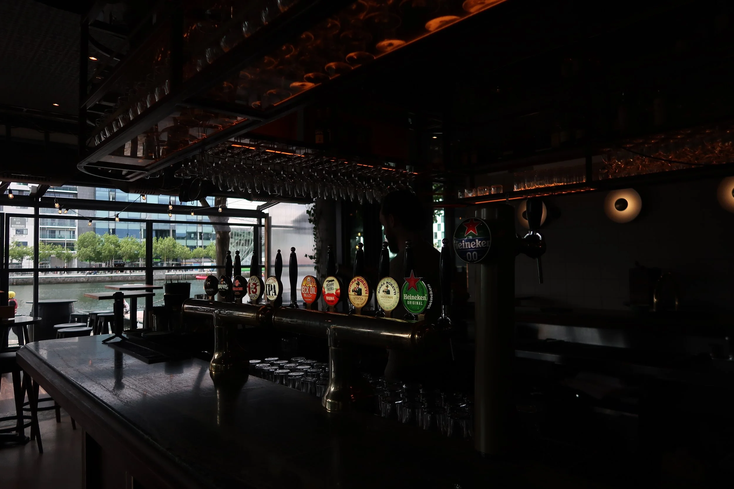 Interior of a bar with a dark ceiling, a row of glassware hanging upside down, and a row of beer taps with labels including Heineken and other brands. Large windows in the background show an outdoor scene with trees and modern buildings.