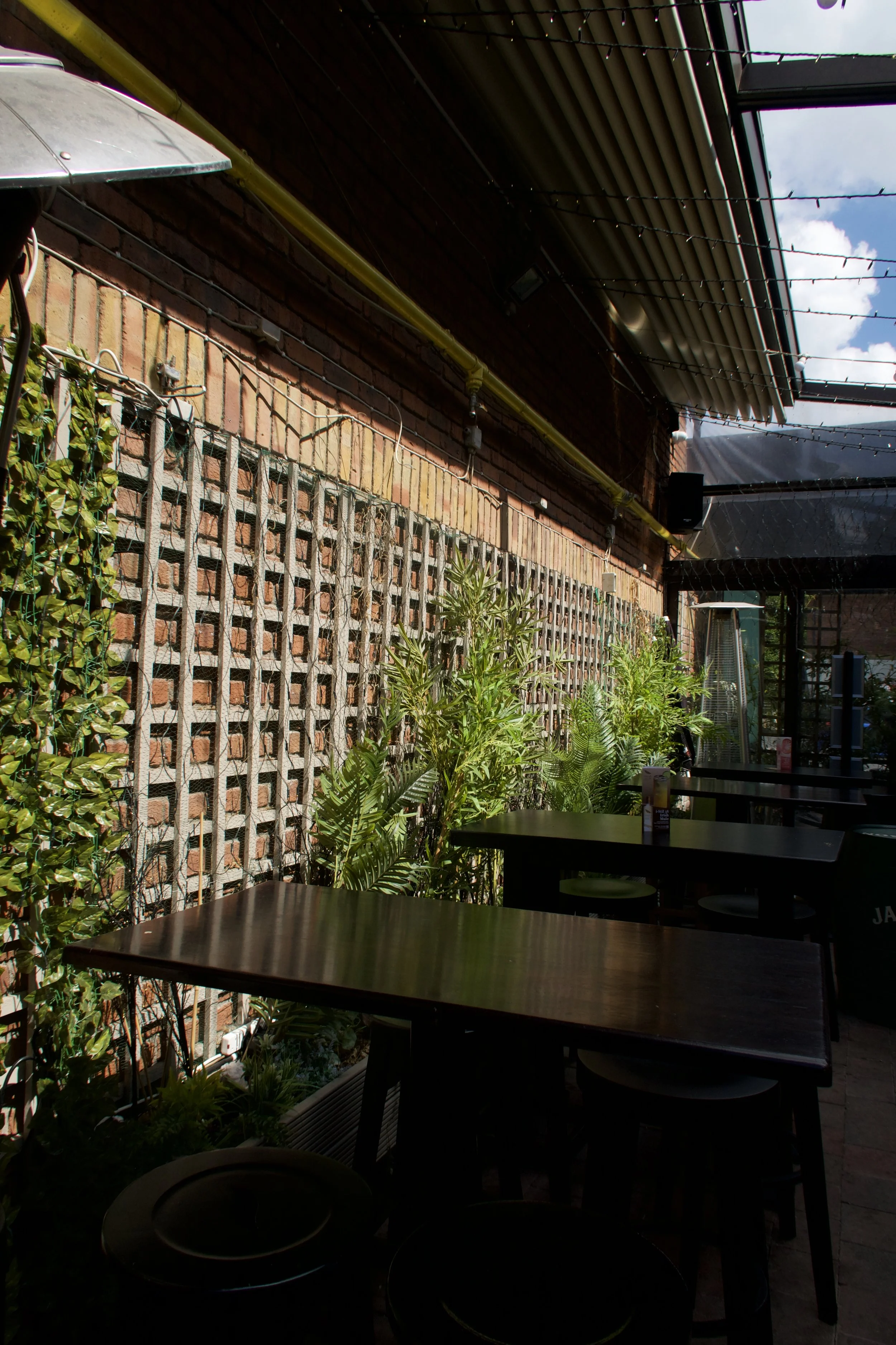 A covered outdoor patio area with tables and chairs, a brick wall with lattice, green plants, string lights, and a partially cloudy sky visible through the roof.