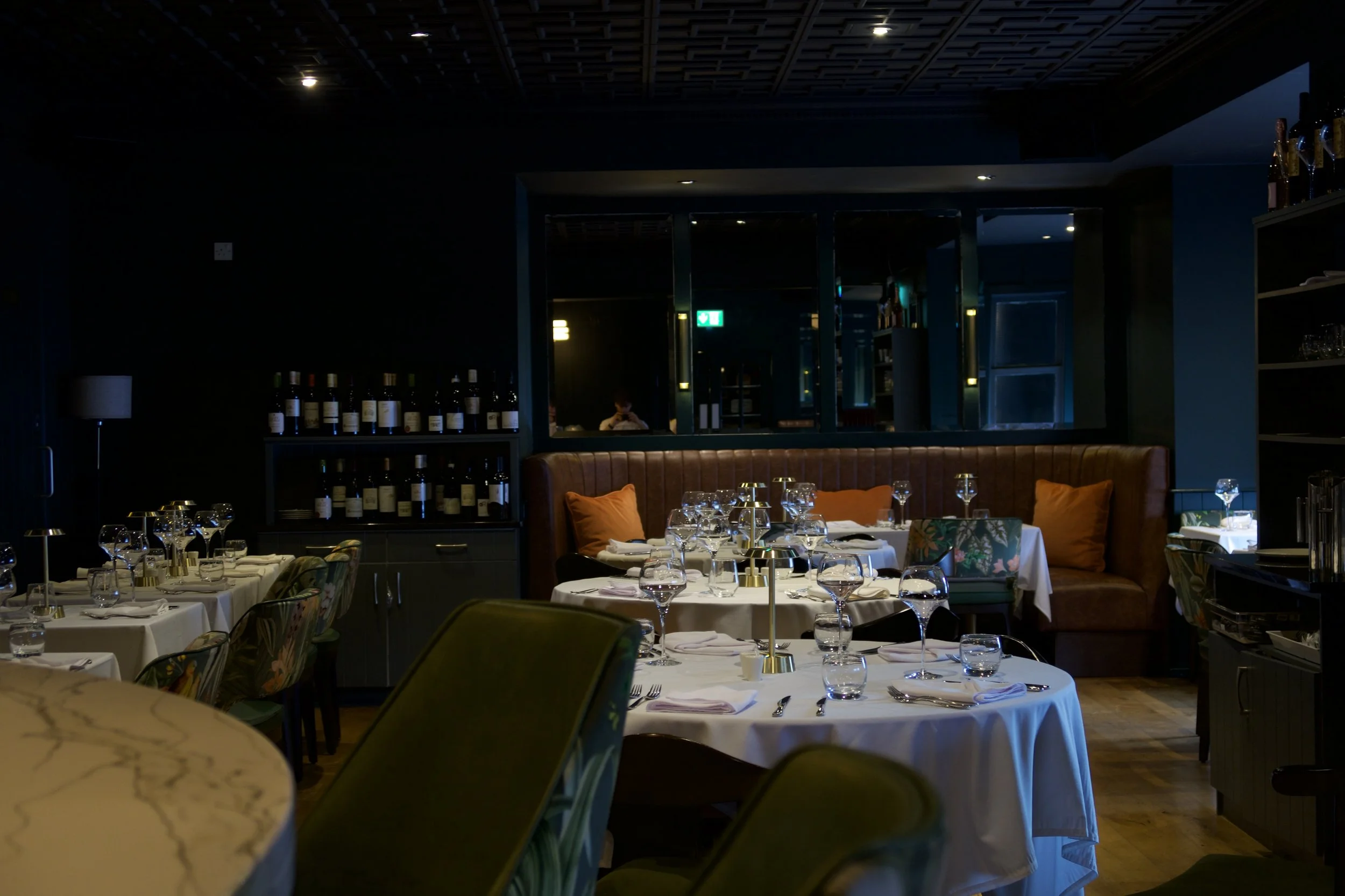 A dimly lit restaurant interior with neatly set tables featuring white tablecloths and glassware, a cushioned seating area with colorful pillows, and shelves of wine bottles against a dark wall. Reflection of a person can be seen in the window.