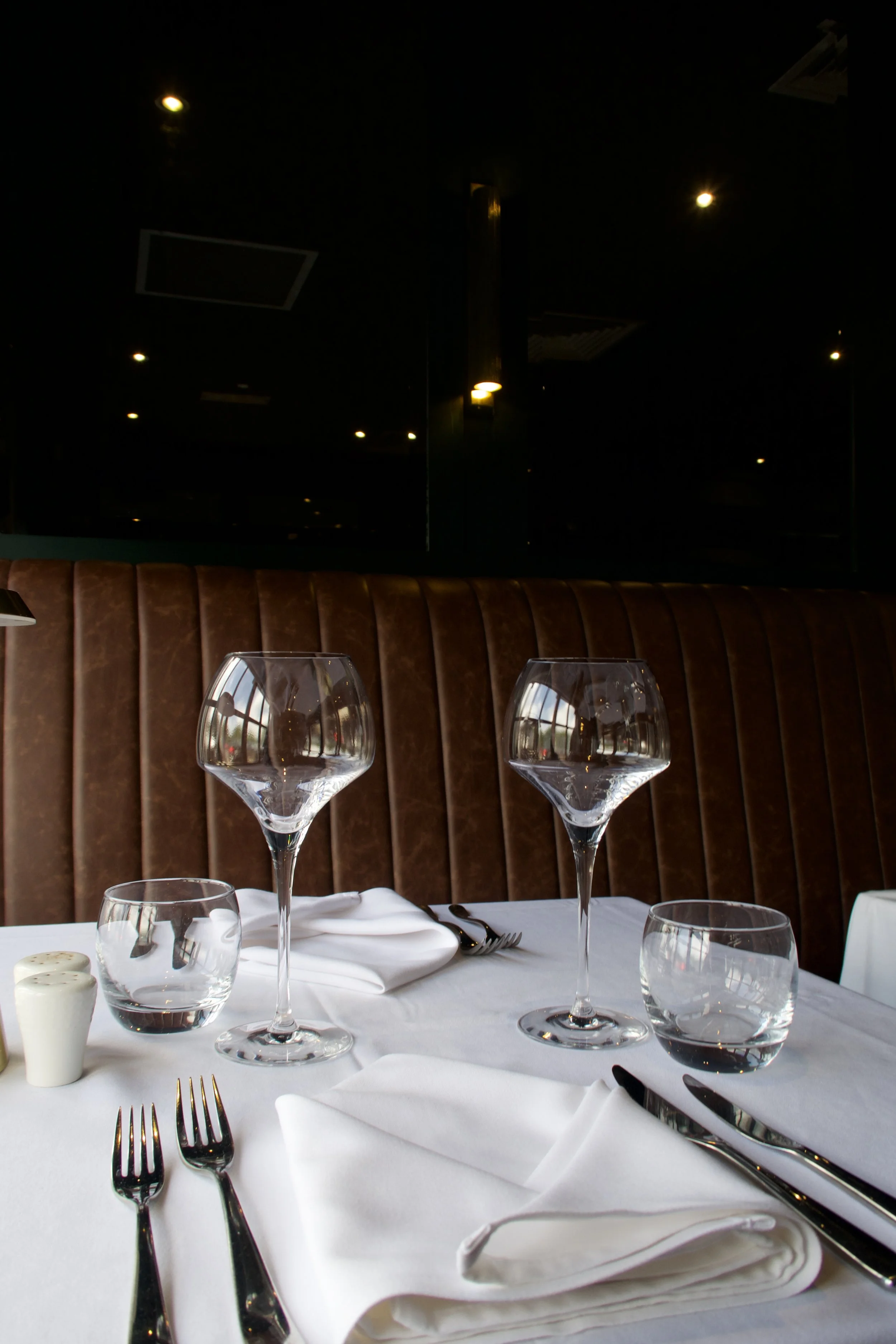 Table setting in a restaurant with wine glasses, water glasses, silverware, white napkins, and a small container of butter. The background has brown upholstered seating and a large window or mirror with yellow lights.