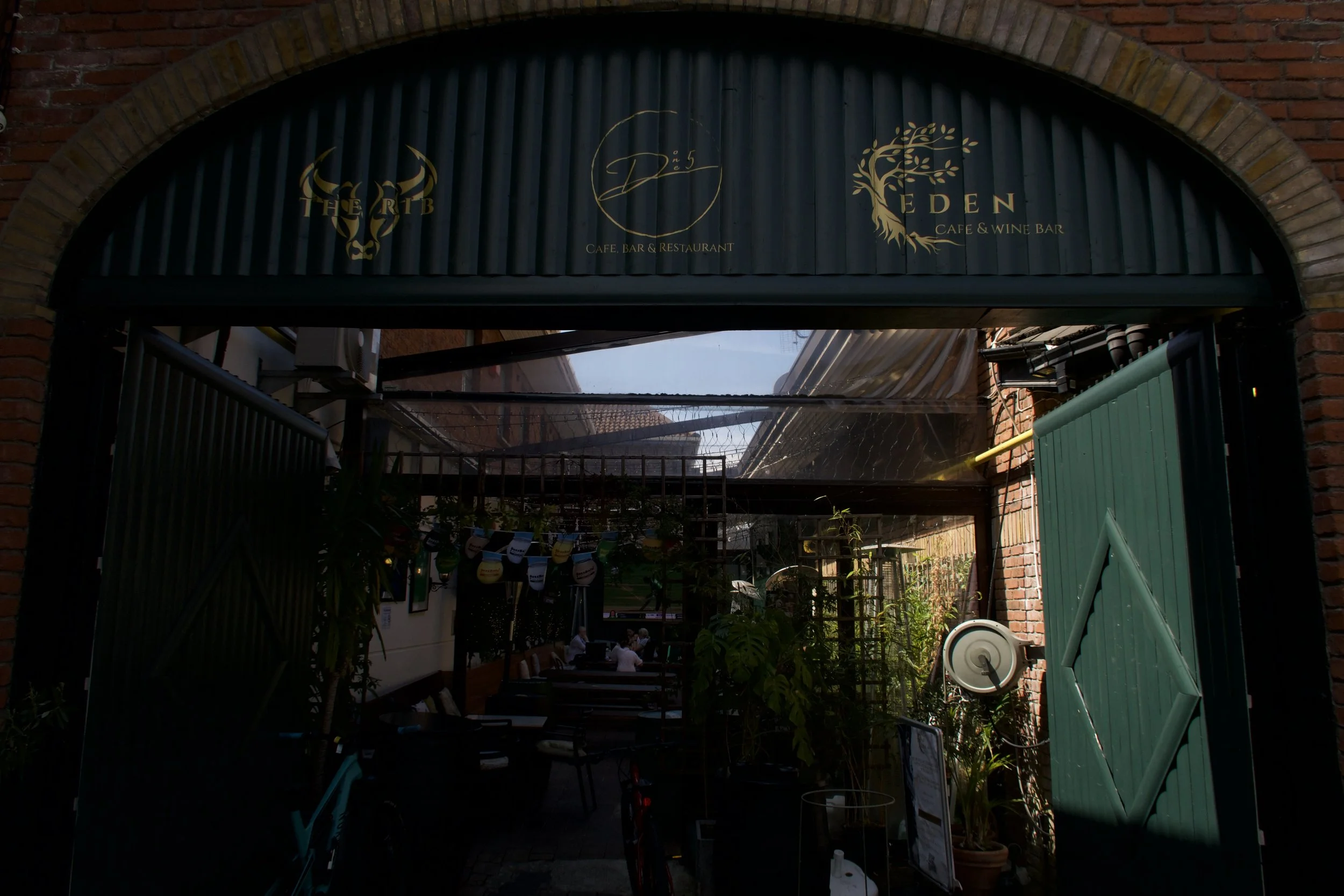 An outdoor restaurant or café with a covered patio area, brick arch and walls, and green gate. Inside, there are tables with group of people dining, and string lights or decorations hanging, with greenery and plants around.