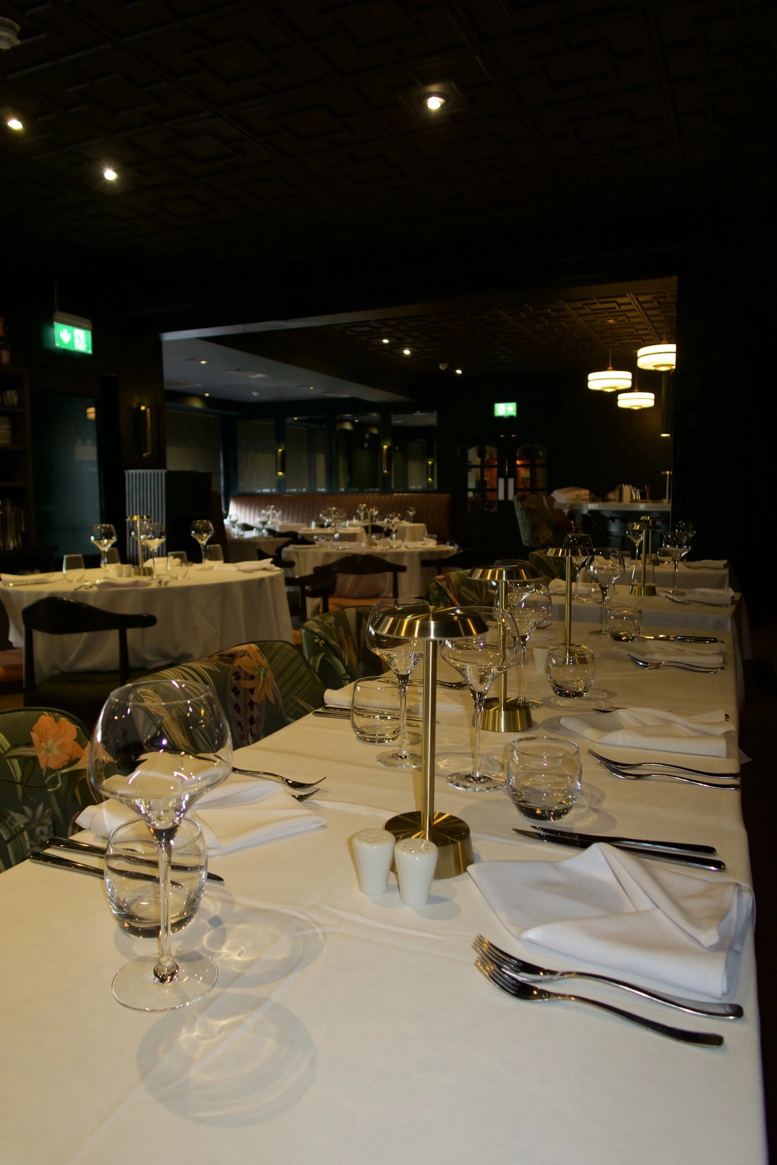 A restaurant dining room with tables covered in white tablecloths, set with wine glasses, plates, silverware, and napkins. The room has ambient lighting, dark walls, and a wooden ceiling, with some modern pendant and recessed lighting fixtures.