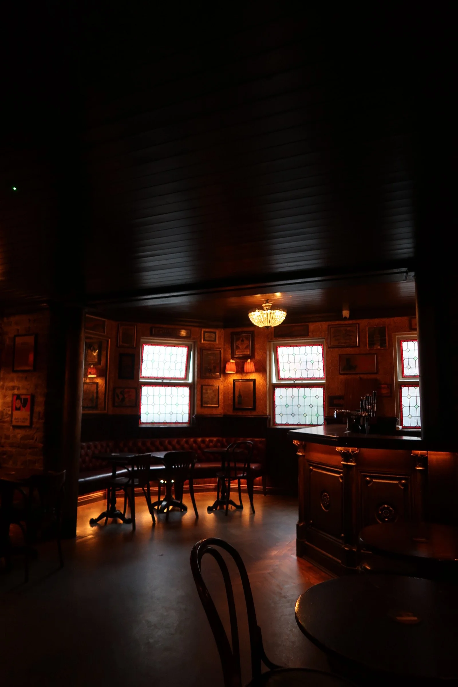 Dimly lit pub or bar interior with dark wood furnishings, a chandelier, framed pictures on the wall, and stained glass windows