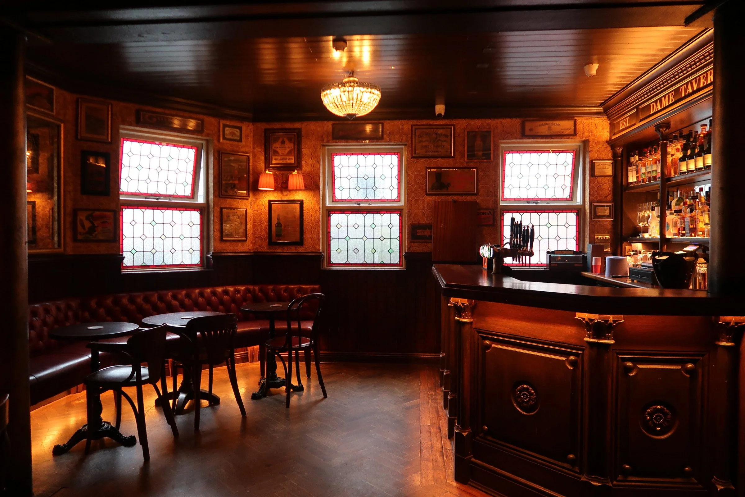 Inside a dimly lit pub with stained glass windows, framed pictures on the walls, a chandelier hanging from the ceiling, a bar with bottles, and tables with chairs.