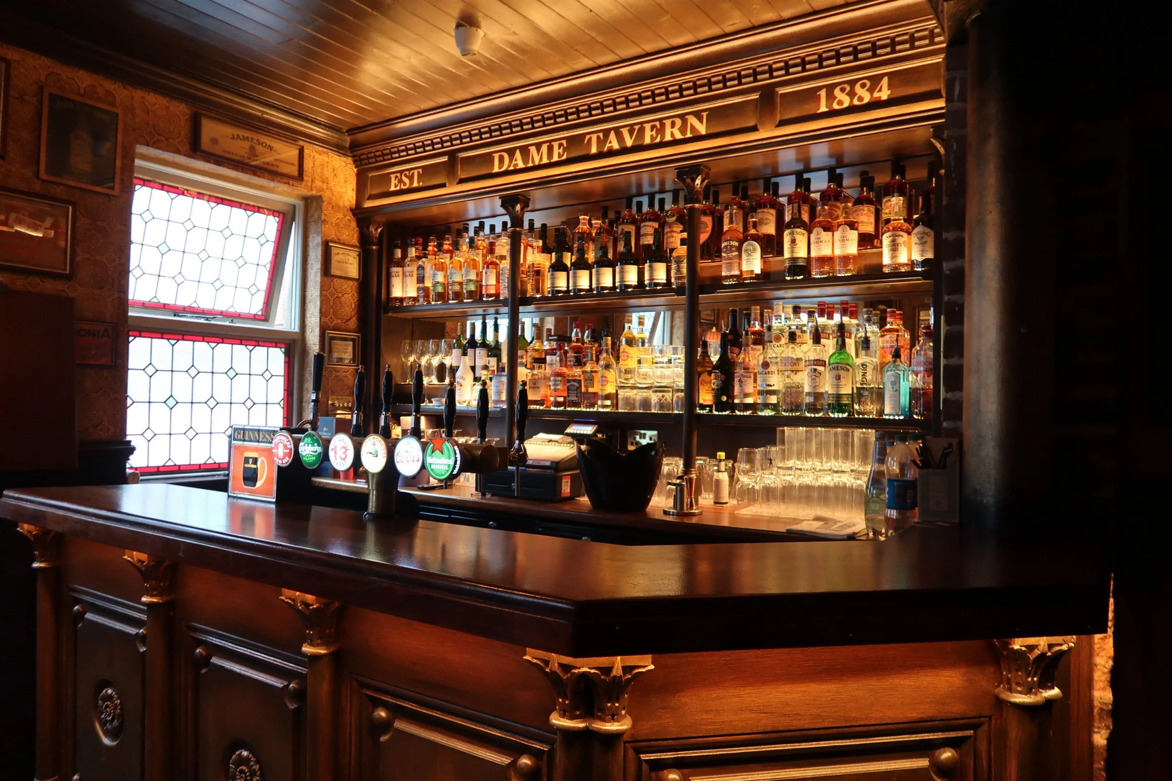 Image of a bar with a dark wooden counter and shelves behind it stocked with various bottles of Jameson Irish whiskey. A row of draft beer taps is visible on the bar, and the establishment's name 'Dame Tavern' with the year '1884' is displayed on a sign above the shelves. There is a stained glass window on the left side, and the interior features warm lighting and vintage decor.