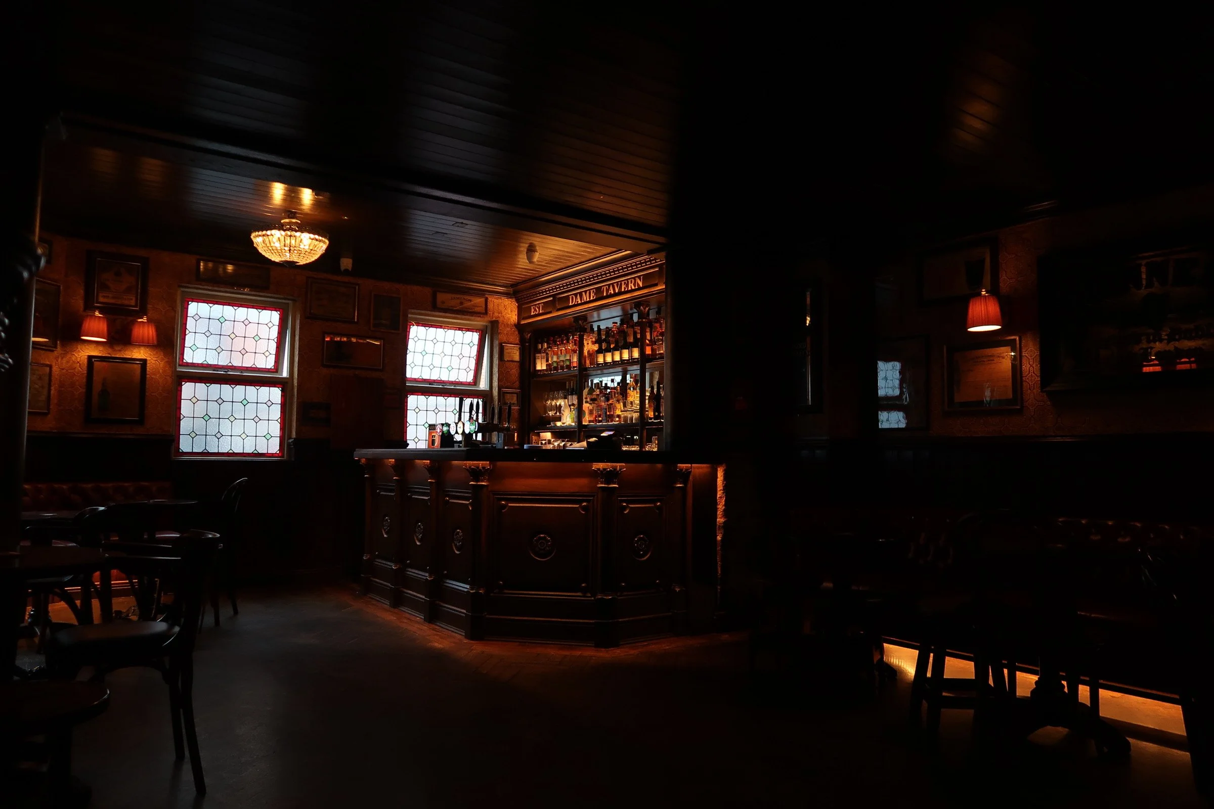 The interior of a dimly lit bar or pub with a wooden bar counter, stained glass windows, framed pictures on the walls, and warm lighting
