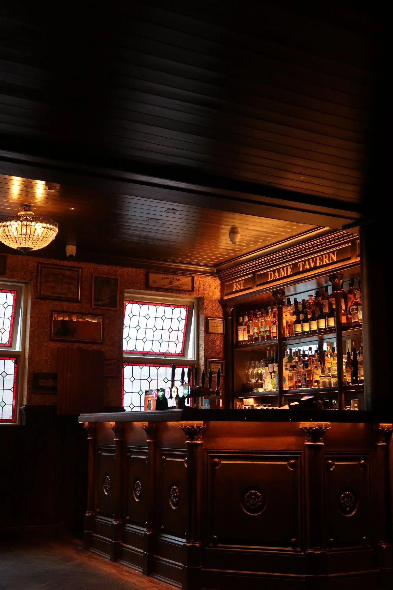 Interior of a vintage-style bar with dark wooden bar counter, bottles on shelves, stained glass windows, and a chandelier lighting fixture.