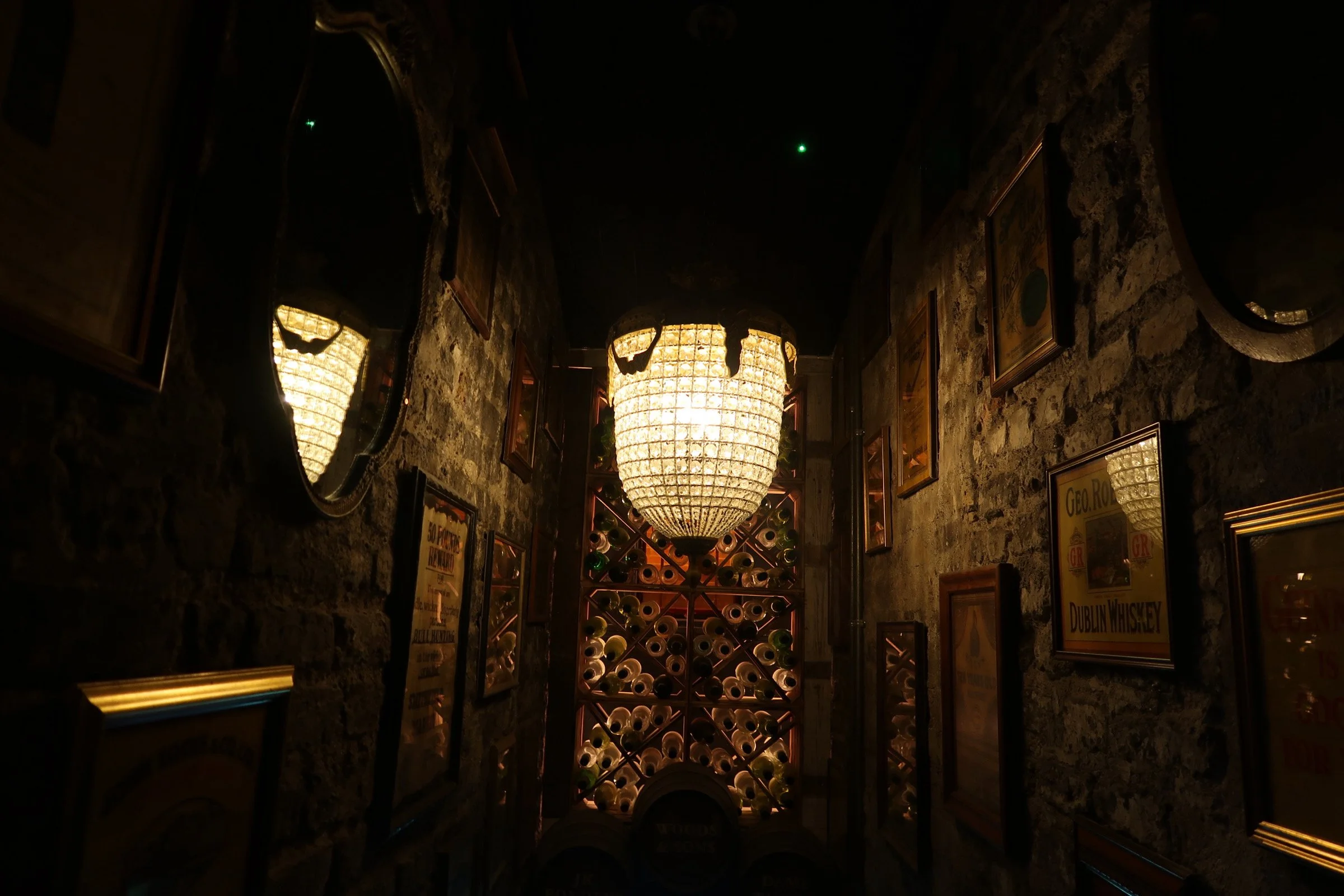 A dimly lit wine cellar with stone walls, a large chandelier, framed posters, and a wine rack filled with bottles.