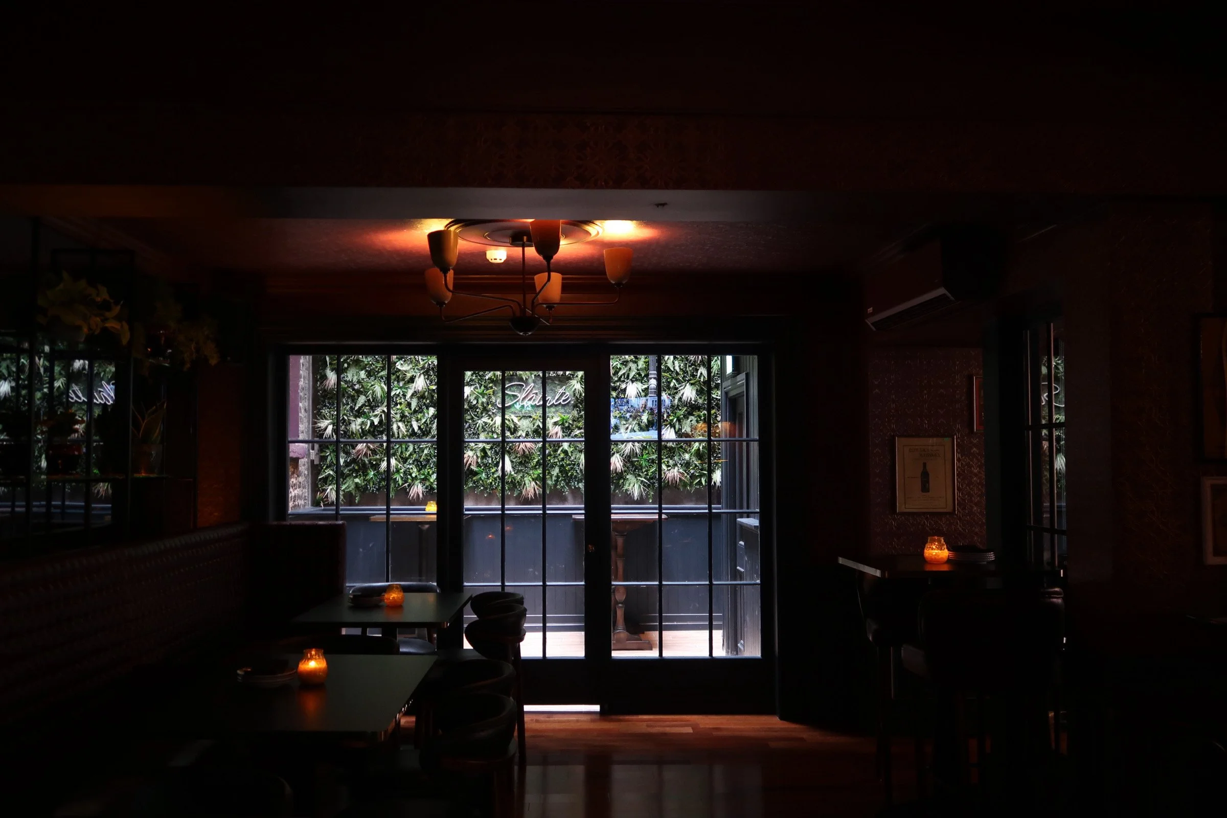 Dimly lit restaurant interior with candles on tables and large windows showing greenery outside.