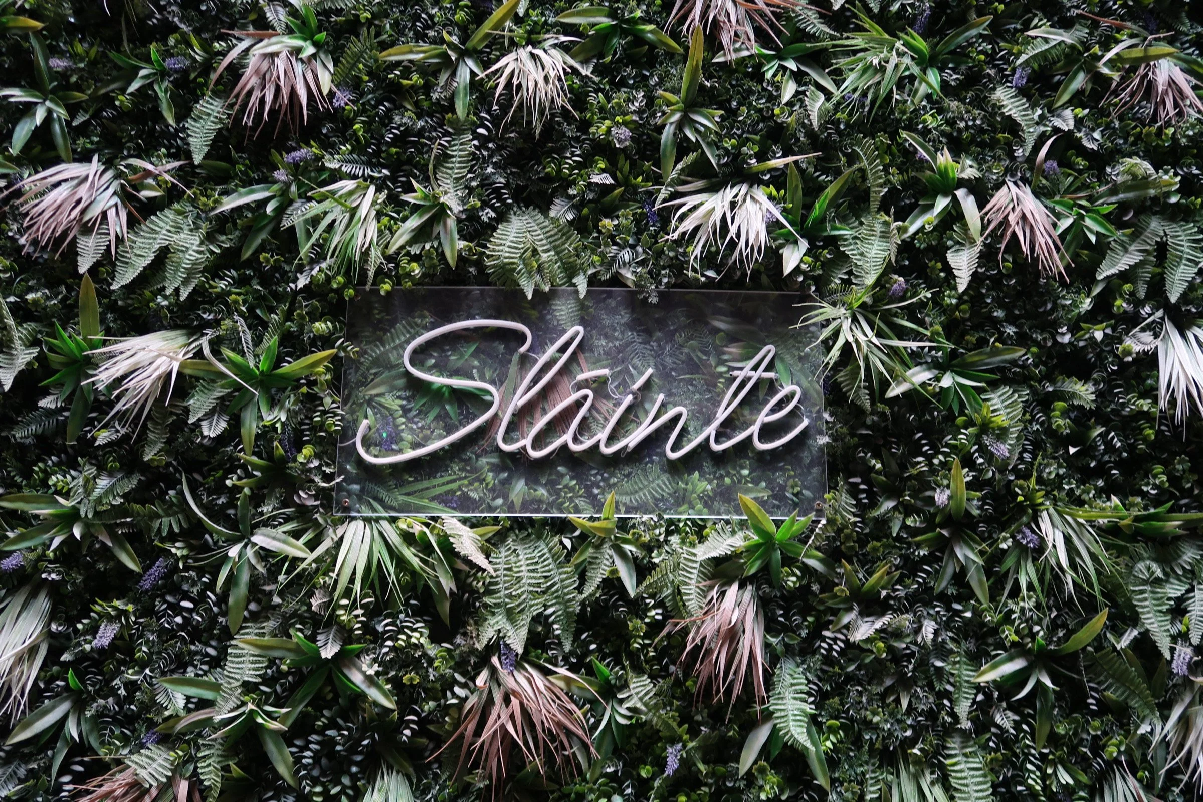 A wall covered in lush green tropical plants with a neon sign that says 'Smile' in the center.