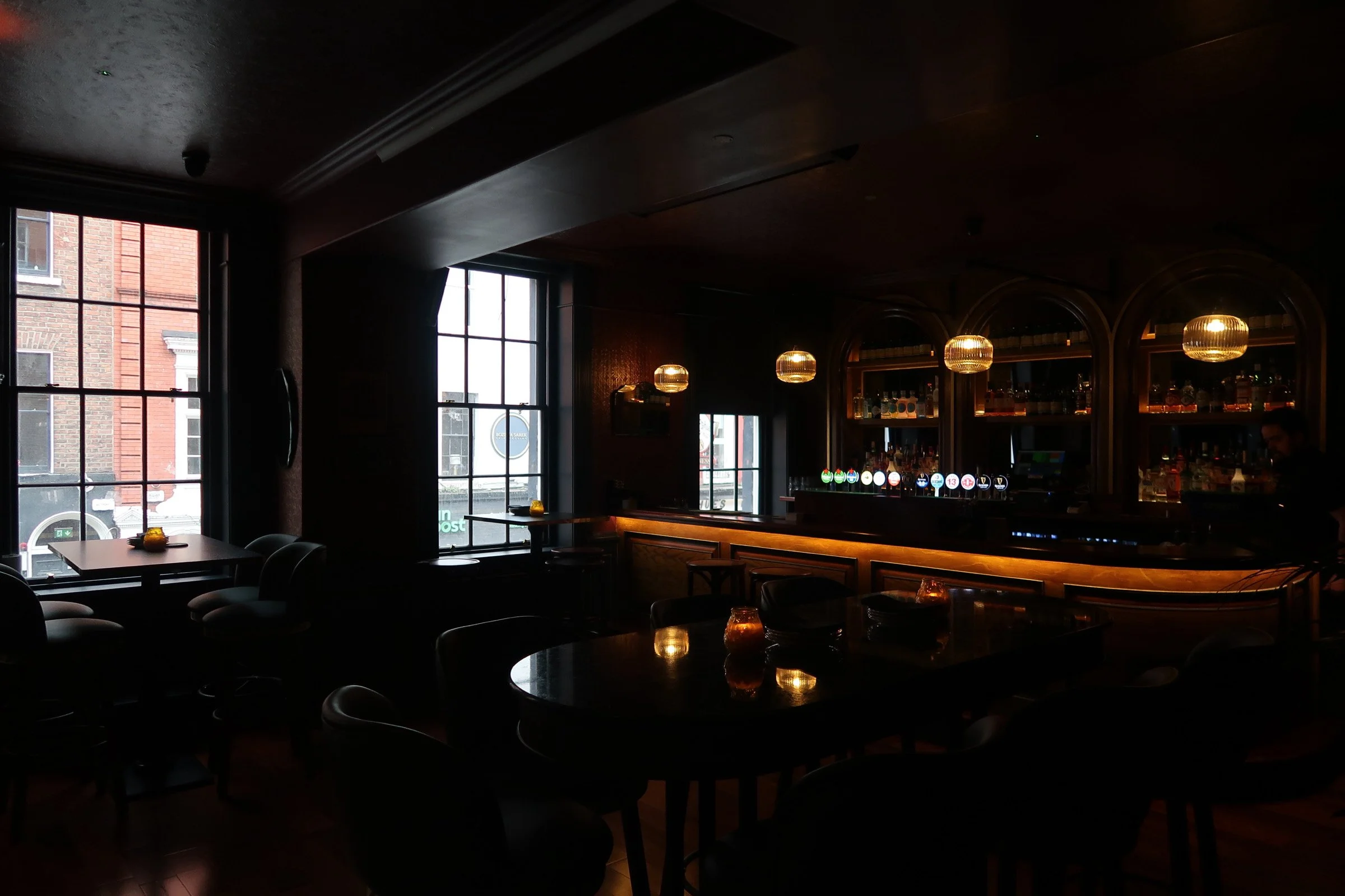 Interior of a dimly lit bar or restaurant featuring large windows, a bar counter with illuminated taps, and hanging pendant lights.