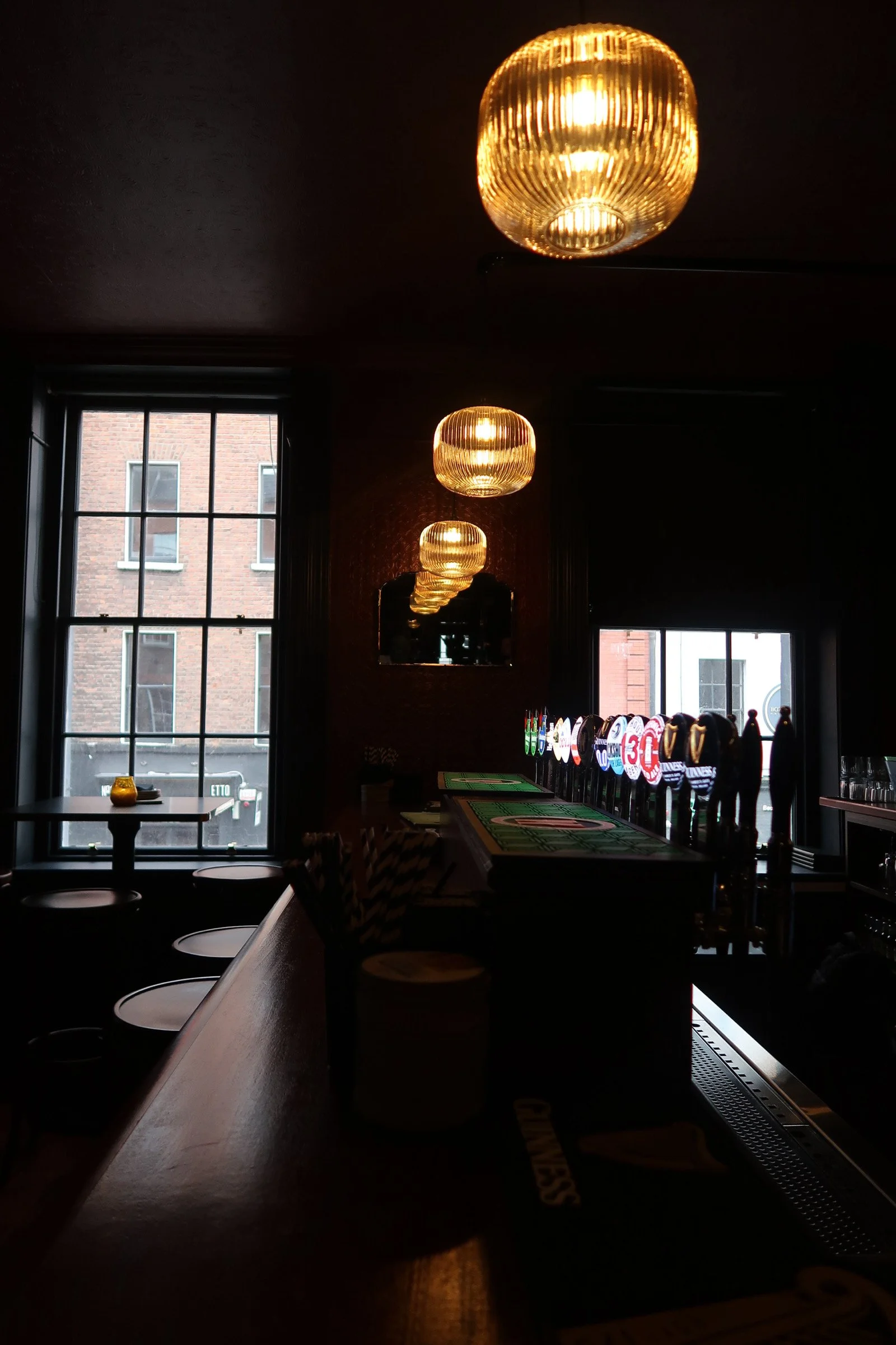Interior of a dimly lit bar with three golden pendant lights hanging from the ceiling and two large windows showing a brick building outside.