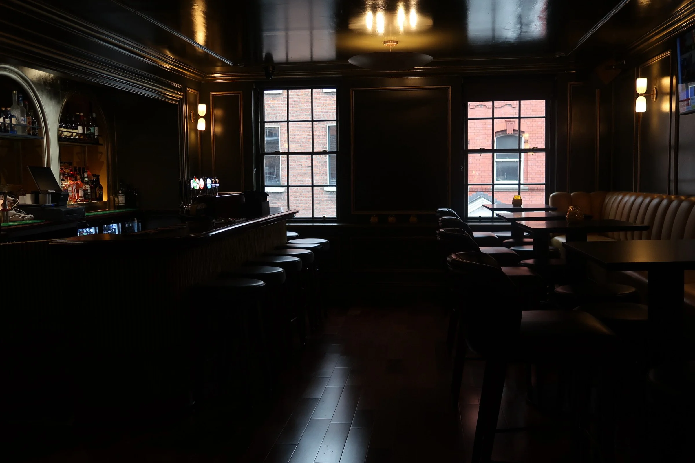 Interior of a dimly lit bar or restaurant with dark walls, large windows showing brick buildings outside, and seating areas including bar stools and tables.