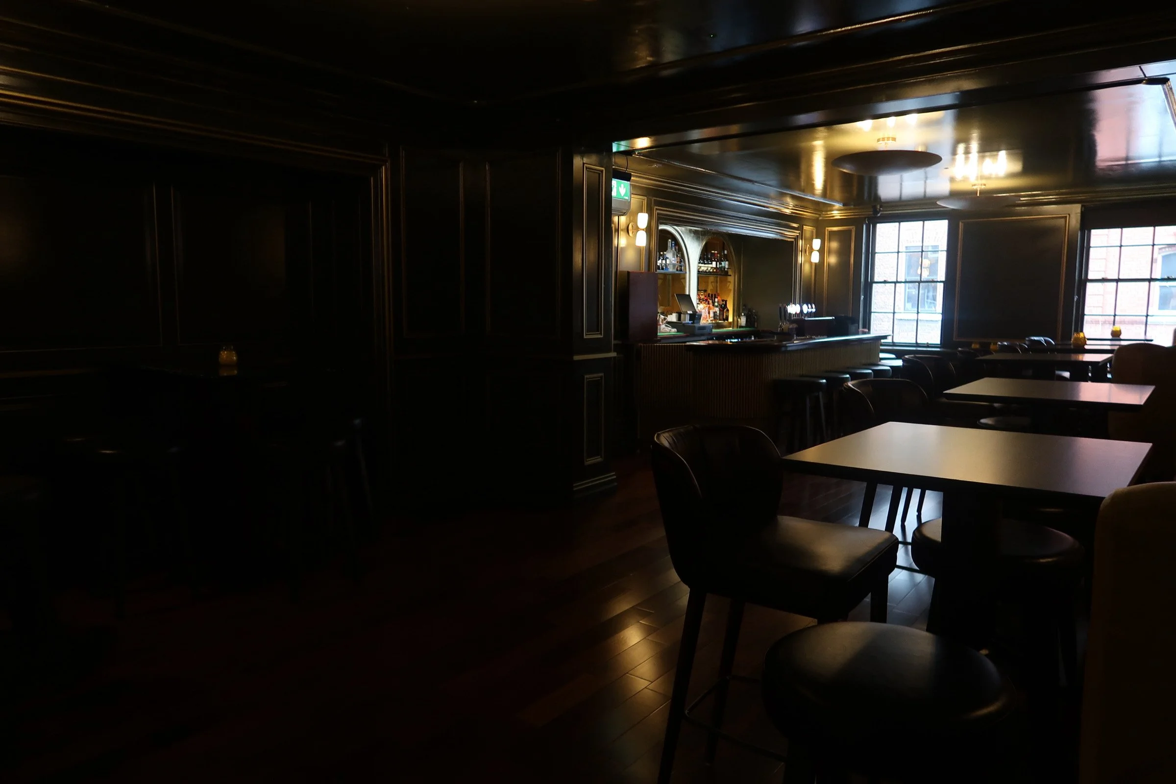 A dimly lit bar area with tables, chairs, and large windows letting in natural light. The bar area has bottles and glasses displayed on shelves, with bar stools in front. The ceiling has decorative lighting, and the overall ambiance is cozy and intimate.