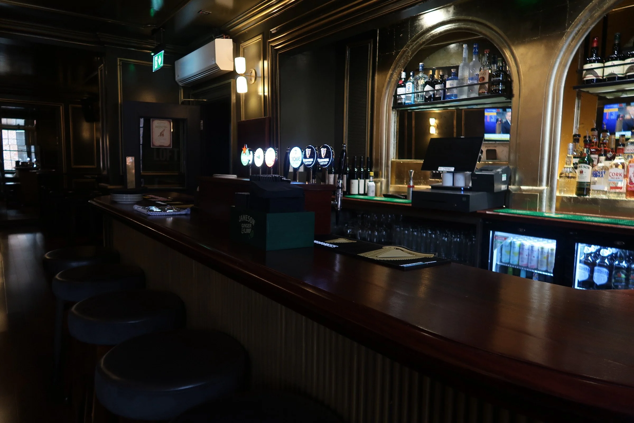 A dimly lit bar with a wooden counter, bar stools, and shelves stocked with various bottles of alcohol. There are TV screens displaying digital content behind the bar.