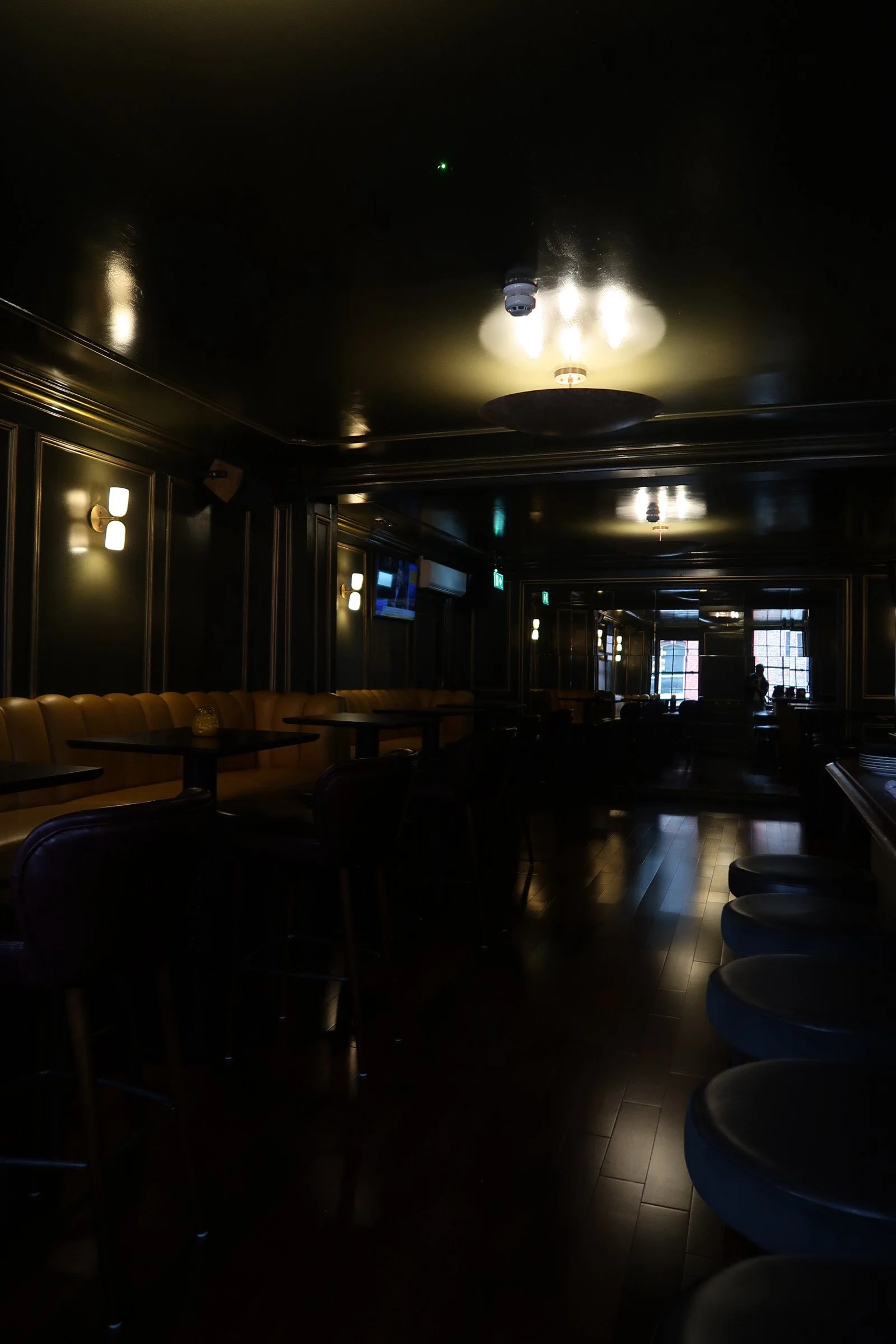 Interior of a dimly lit restaurant or bar with black walls, gold accents, and modern lighting fixtures. There are yellow booths along the wall and chairs around tables, with some windows visible in the background.