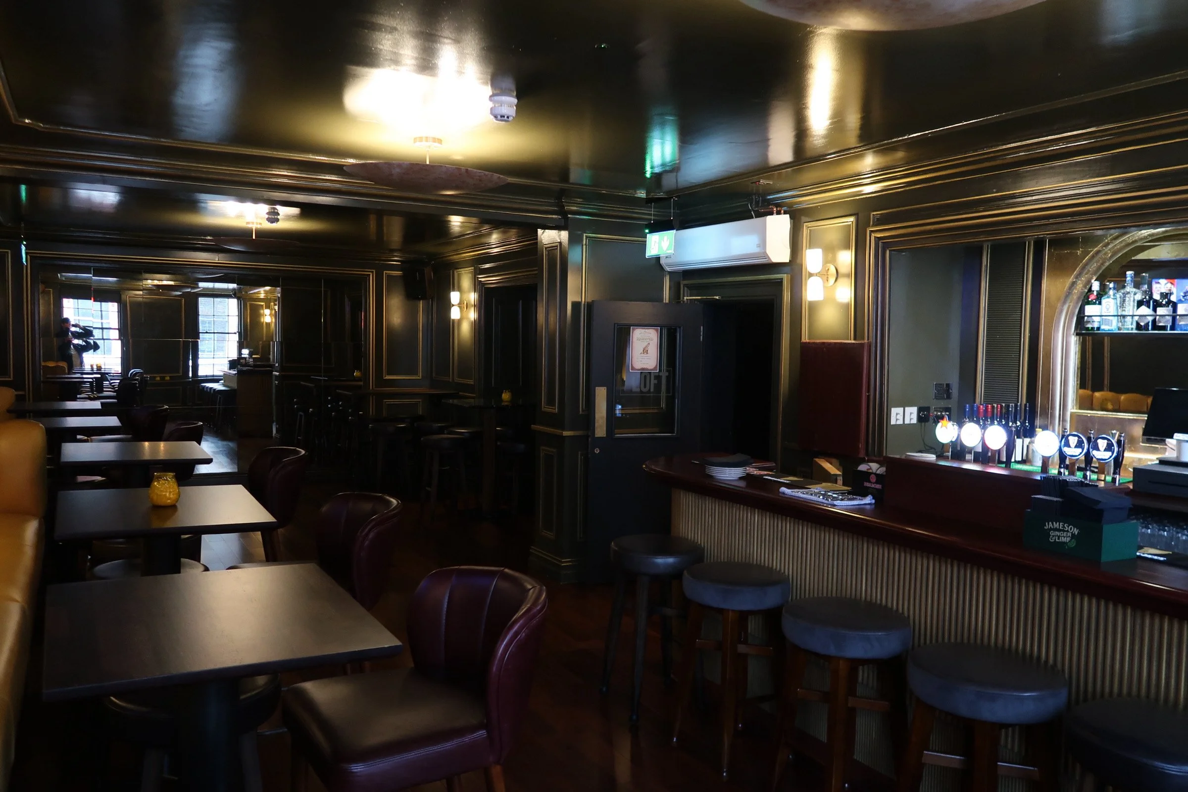 Interior of a dimly lit bar or restaurant with dark walls and gold trim. There are tables and chairs, a bar with various bottles, and a person taking a photo near a window.