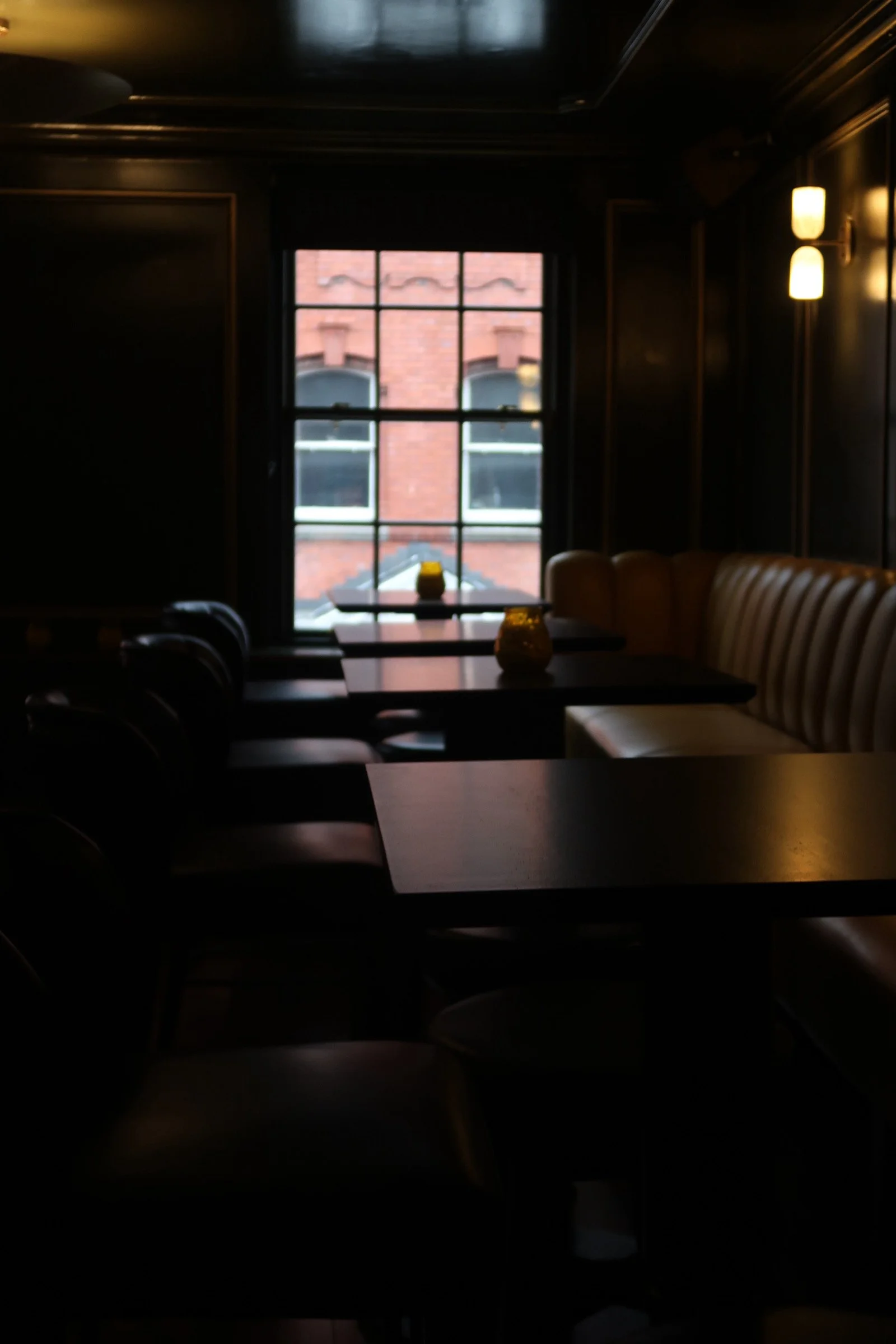 A dimly lit interior of a restaurant or cafe with wooden walls, a window showing a brick building outside, a booth with a beige cushioned bench, and a table with yellow candles.