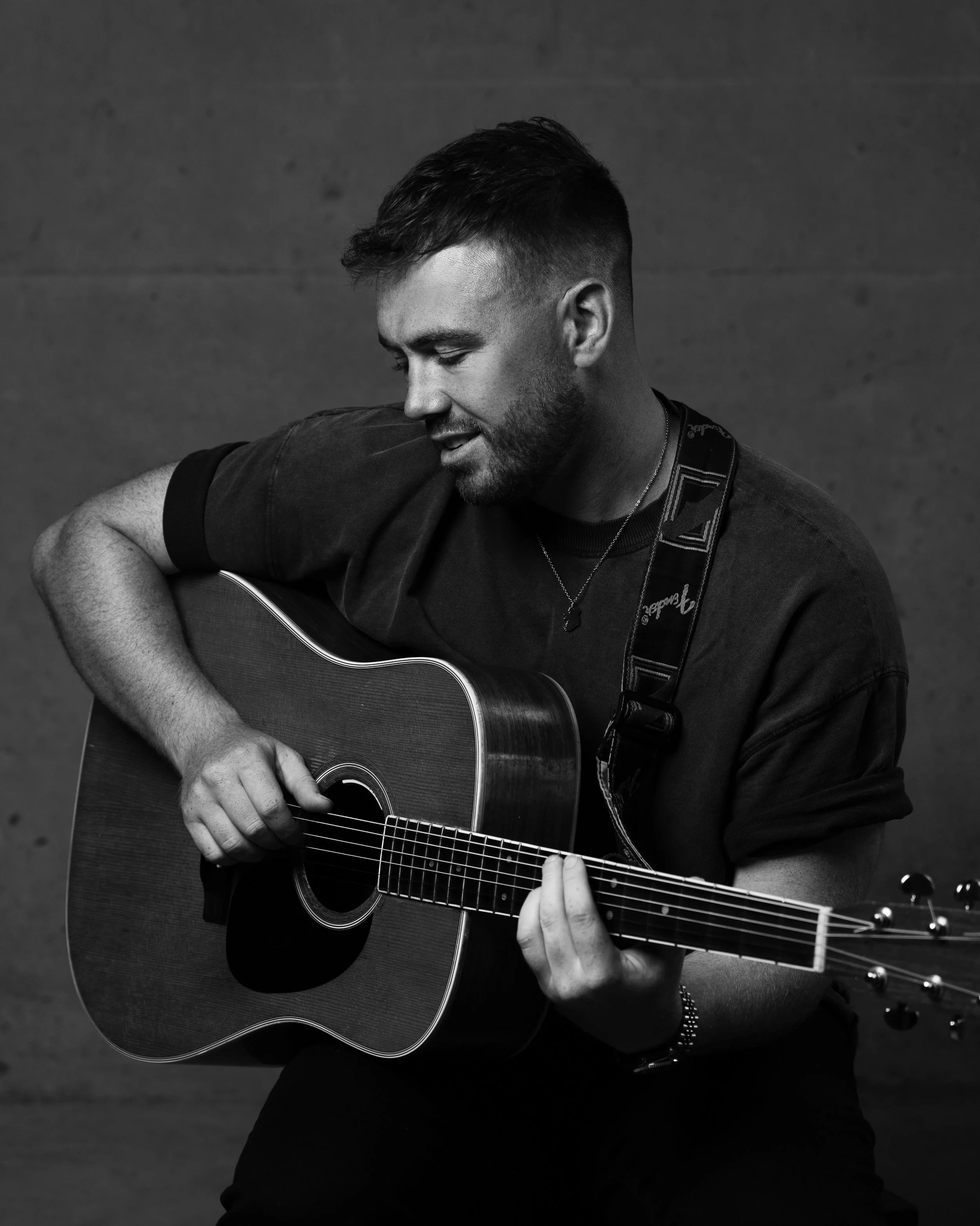 Black and white photo of a man with short hair and a beard playing an acoustic guitar, with a neutral background.