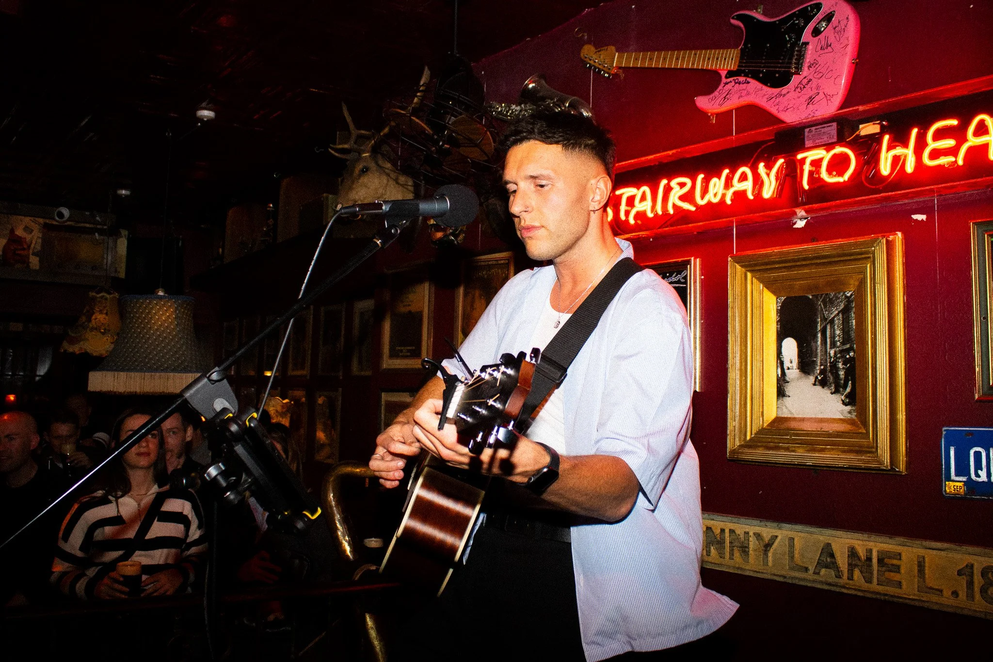 A man playing an acoustic guitar in a bar, with a neon sign that reads 'Fairway to HELL' behind him, and a pink electric guitar hanging on the wall.