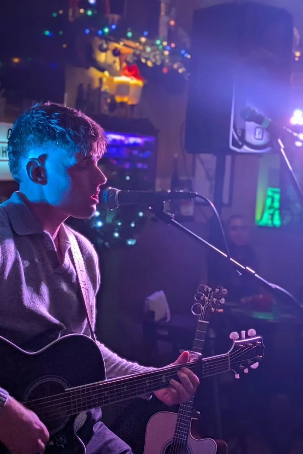 Young male musician singing into a microphone while playing an acoustic guitar during a live performance in a dimly lit venue with colorful lights.