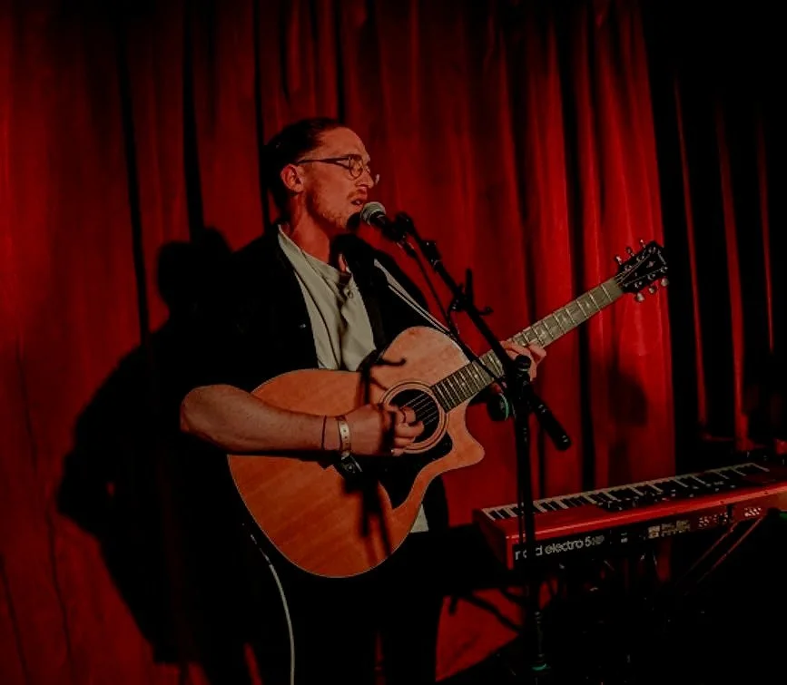 A man playing an acoustic guitar and singing into a microphone on stage with red curtains in the background.