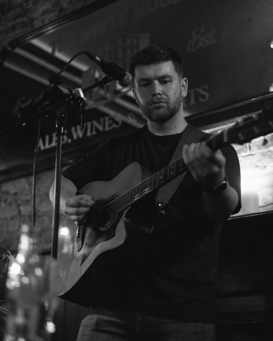 A man playing an acoustic guitar on stage at a venue with a brick wall background, black and white photo.