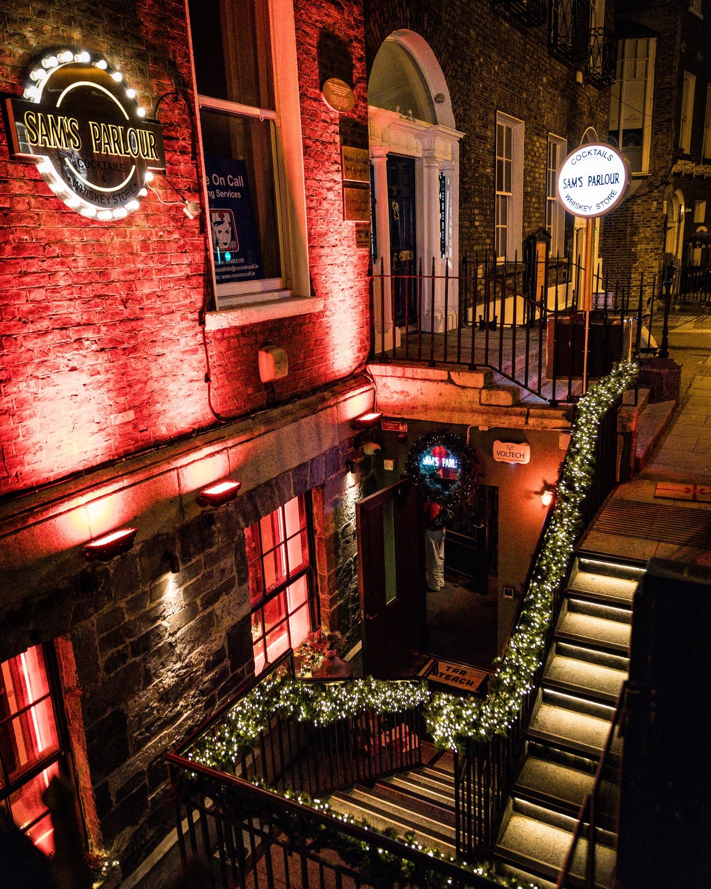 Night scene of a brick building with illuminated signs for Sam's Parlour, a whiskey store and cocktail bar, decorated with festive holiday lights on the staircase railing and around the entrance, with some windows lit from inside.