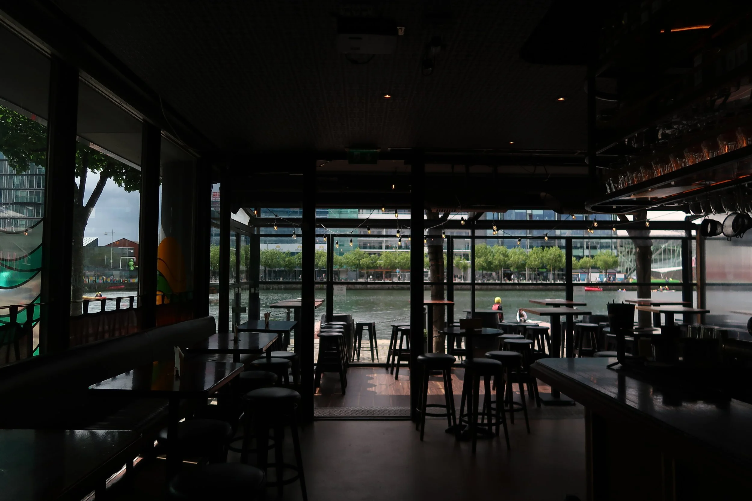 Interior of a dimly lit restaurant or cafe with large windows overlooking a river or canal, trees, and buildings outside; restaurant furniture including tables and stools.