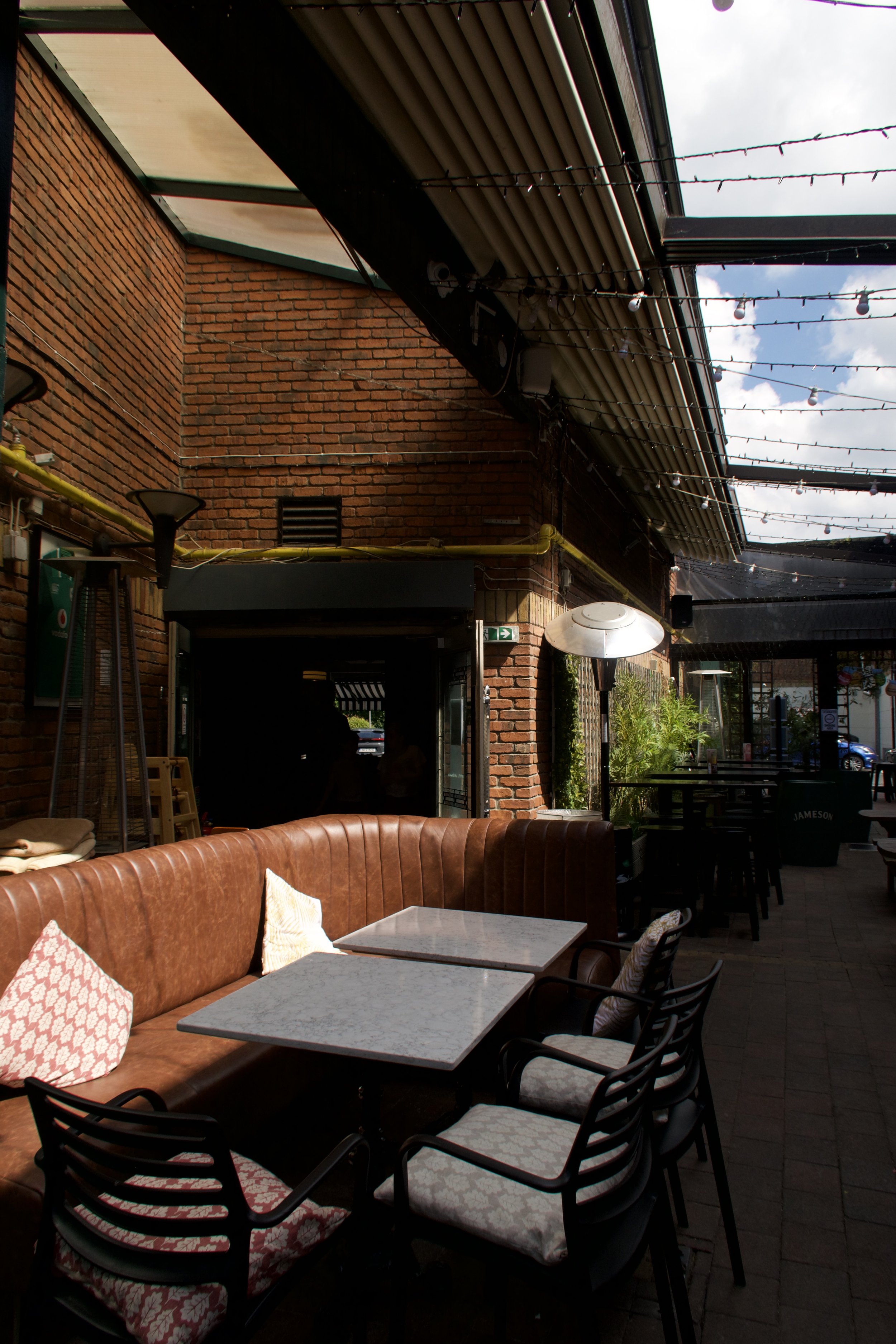 Outdoor patio area with brick walls, string lights overhead, and a mix of seating including a leather sofa with patterned cushions, and black chairs around tables. Part of the sky with clouds is visible through the open roof structure.