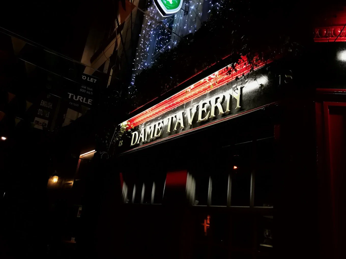 Night view of a bar or restaurant with illuminated sign saying 'Dame Taverna' and decorative string lights.