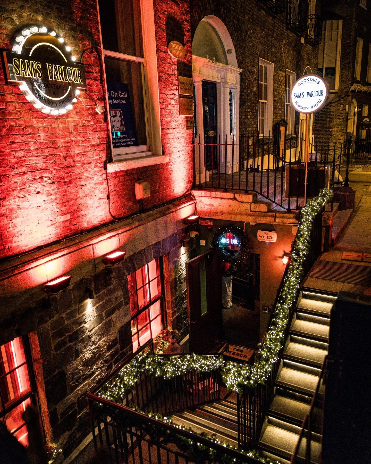 Night scene of a brick building with illuminated signs for Sam's Parlour, a whiskey store and cocktail bar, decorated with festive holiday lights on the staircase railing and around the entrance, with some windows lit from inside.