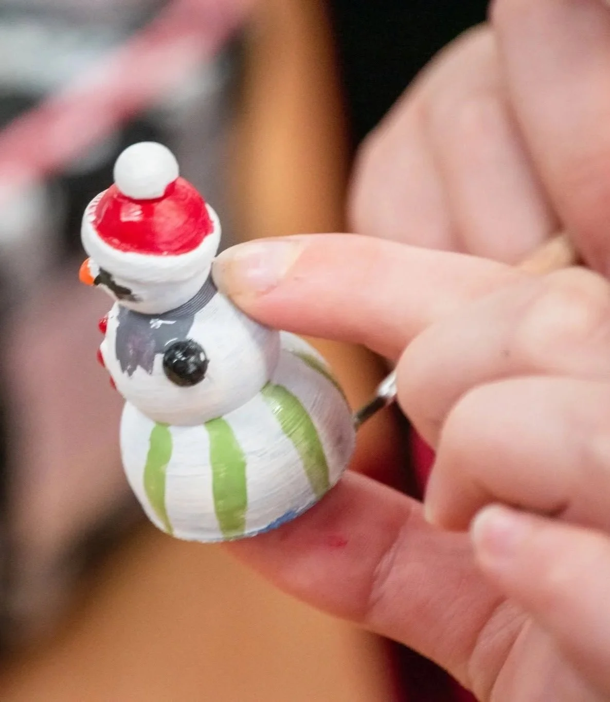 A close-up of a small snowman ornament with a red Santa hat, painted details, and green stripes on its base, held between someone's fingers.