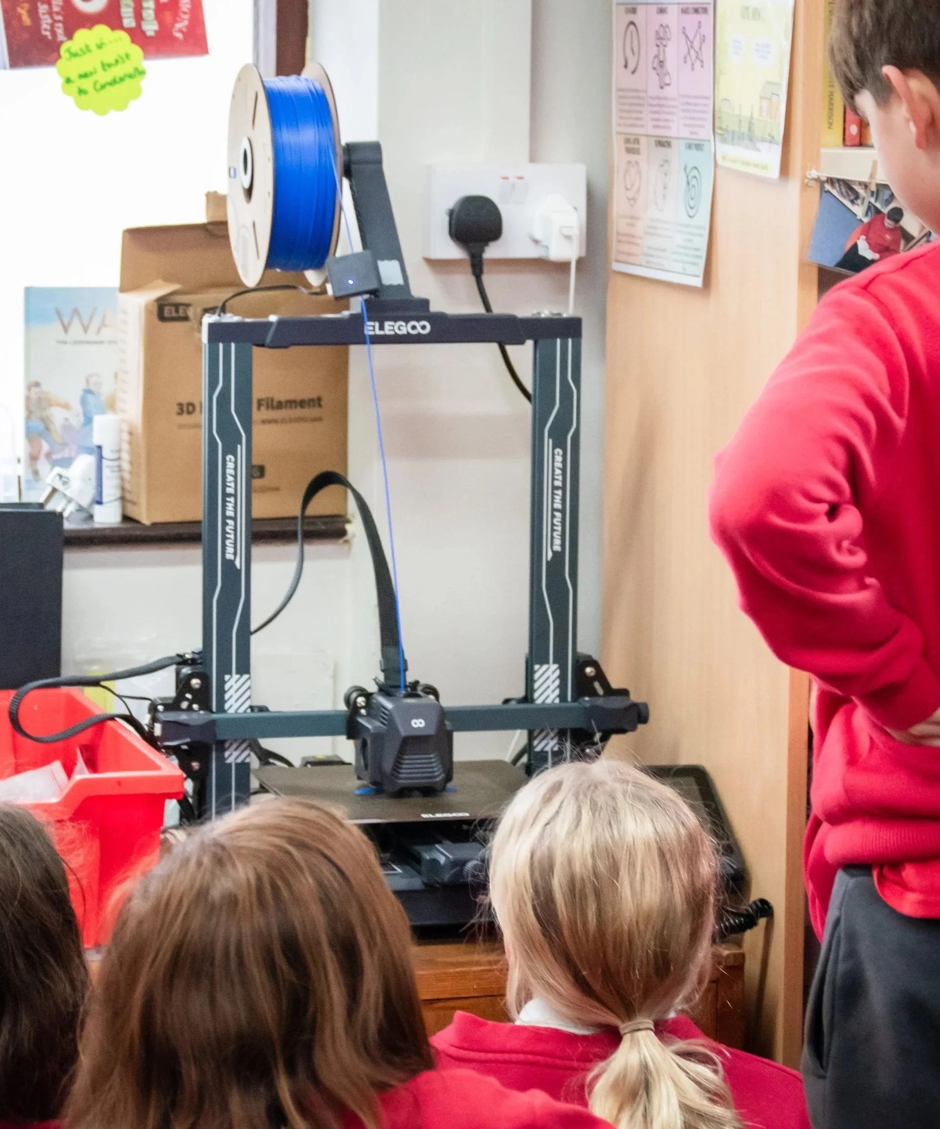 Students gathered around a 3D printer in a classroom, observing it in operation.