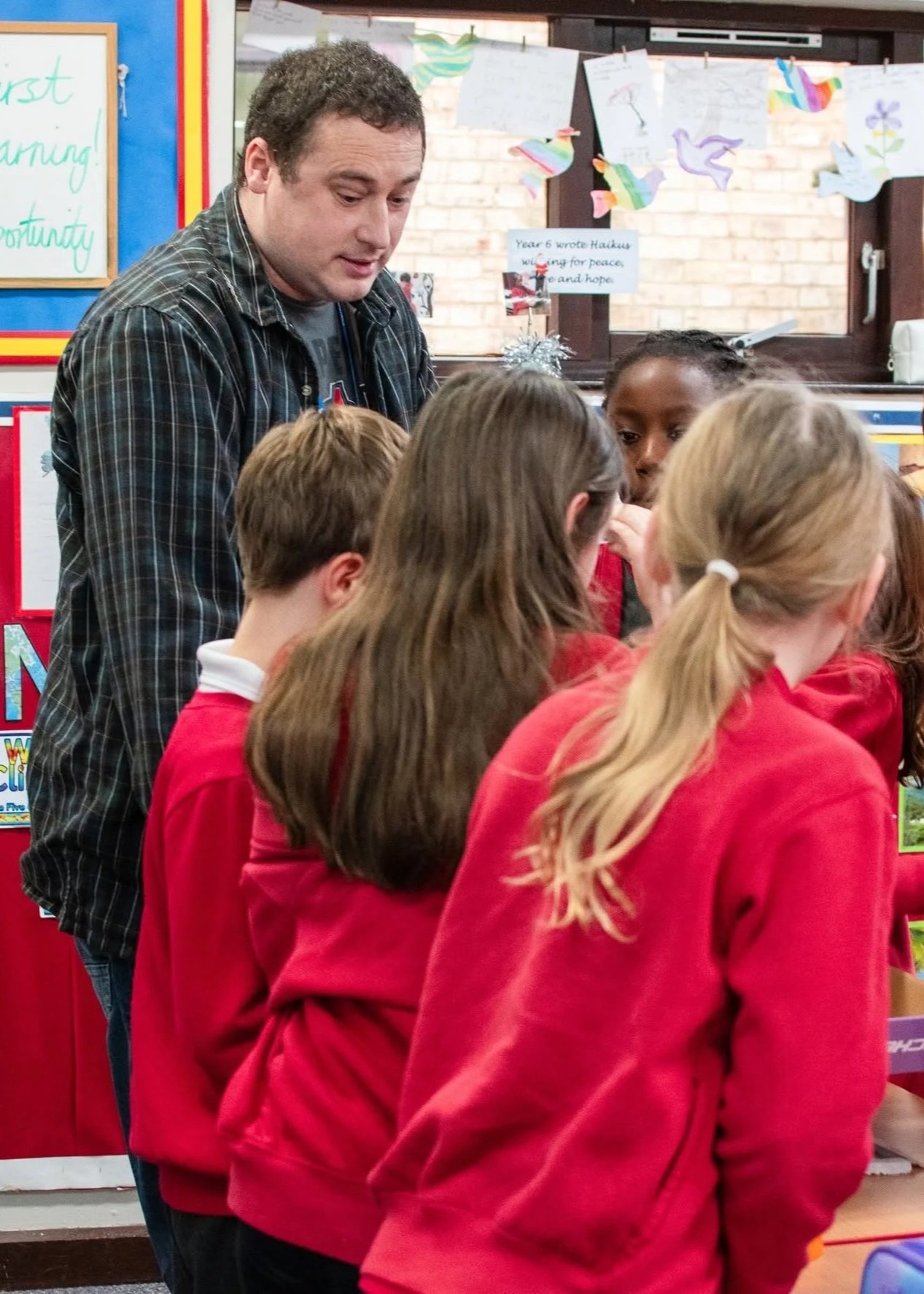 A male teacher engaging with a group of students with their backs to the camera in a classroom, decorated with colorful paper crafts on the windows and educational posters on the wall.