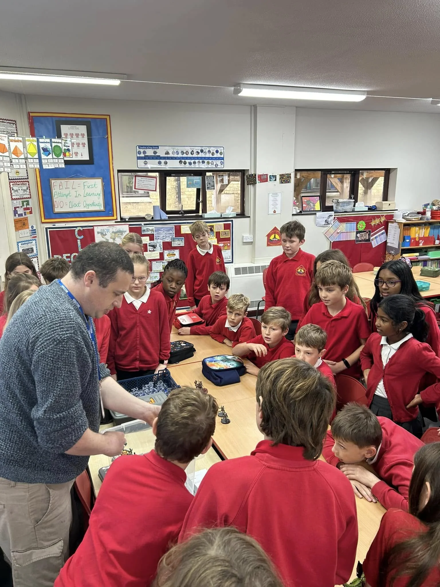 A classroom full of students wearing red uniforms gathered around a table, with a teacher engaged in an activity. The classroom has educational posters and decorations on the walls.