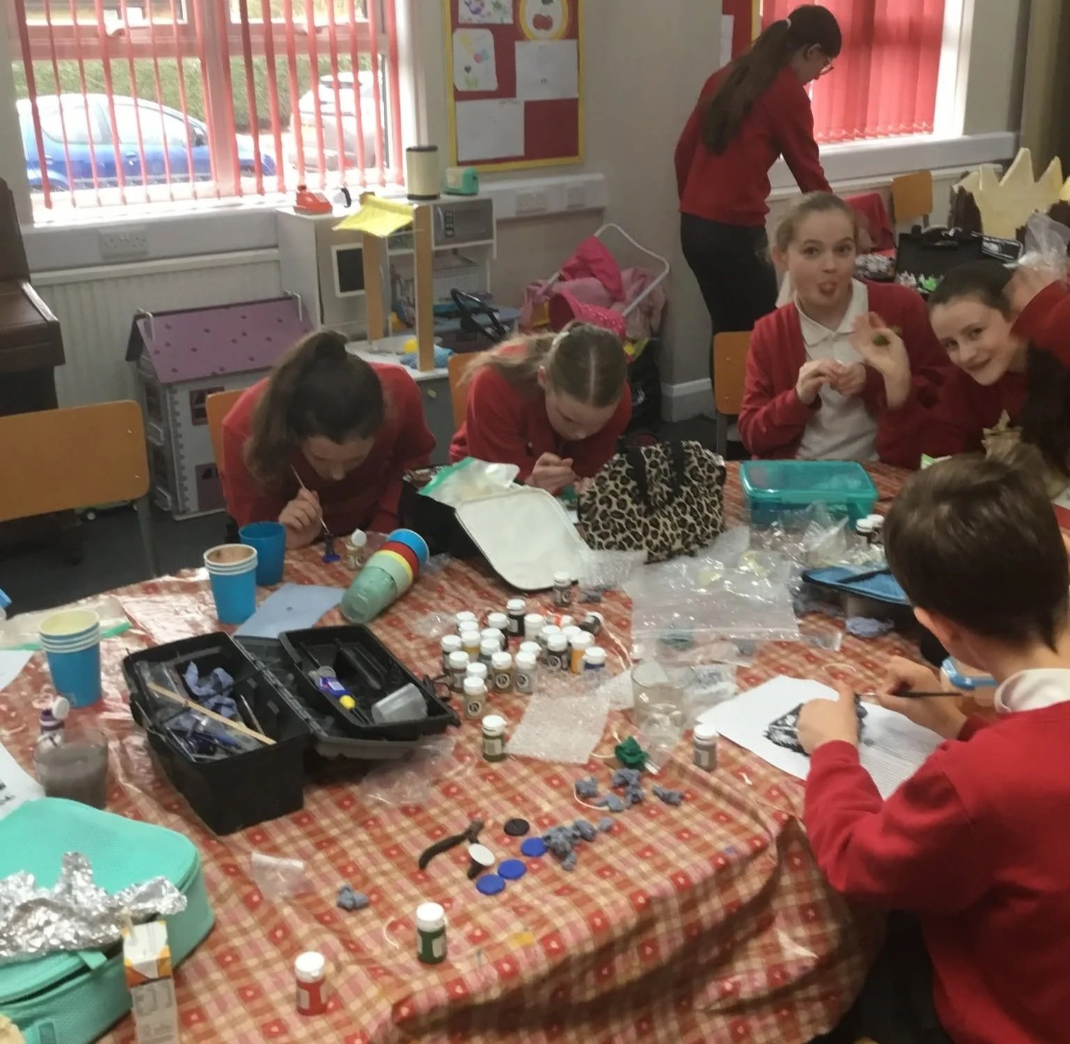 Children in red school uniforms painting and crafting at a table with art supplies, including paint bottles, paper, and containers, in a classroom.