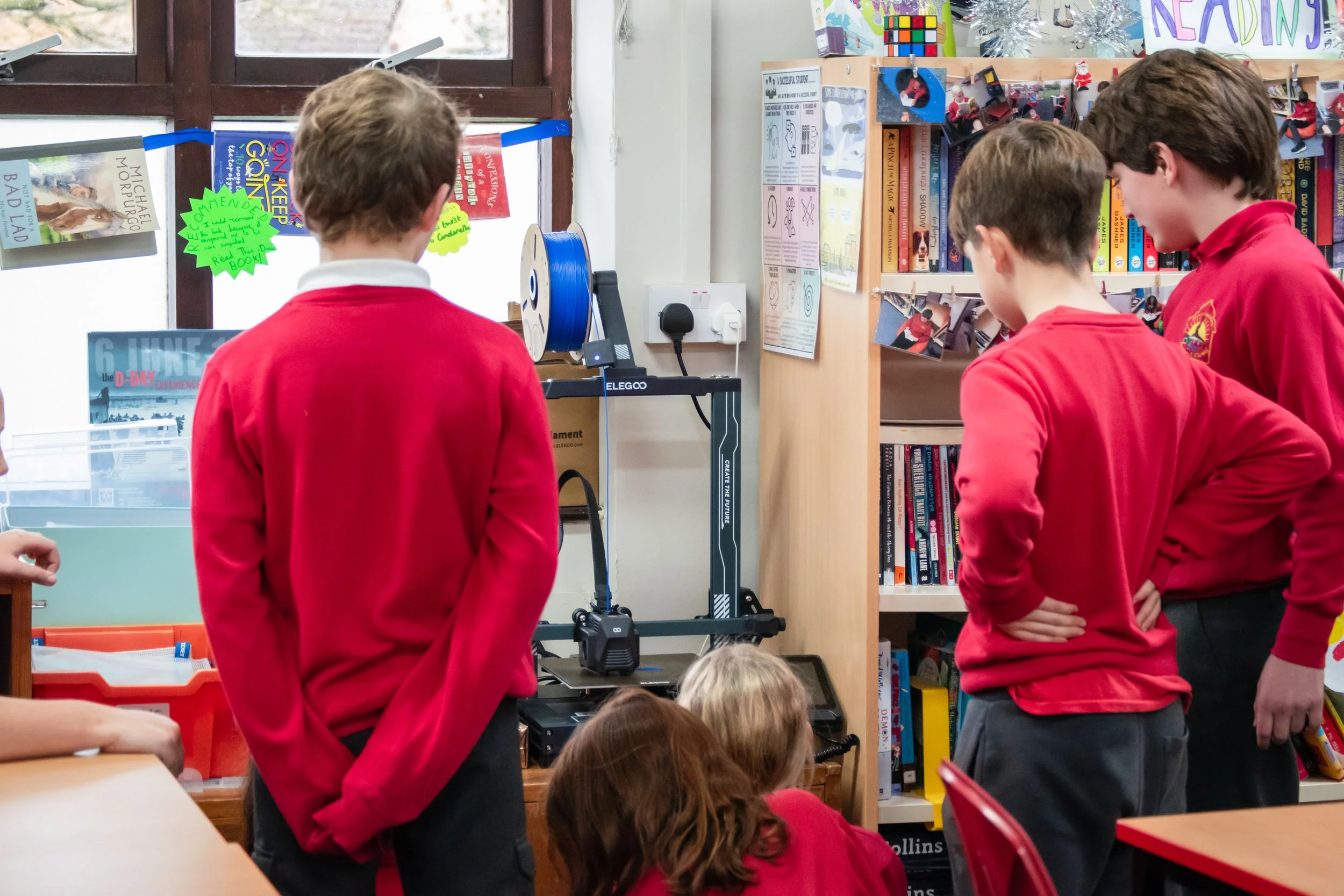 Three boys in red school uniforms standing in a classroom, observing a science project with a 3D printer, surrounded by books and classroom decorations.