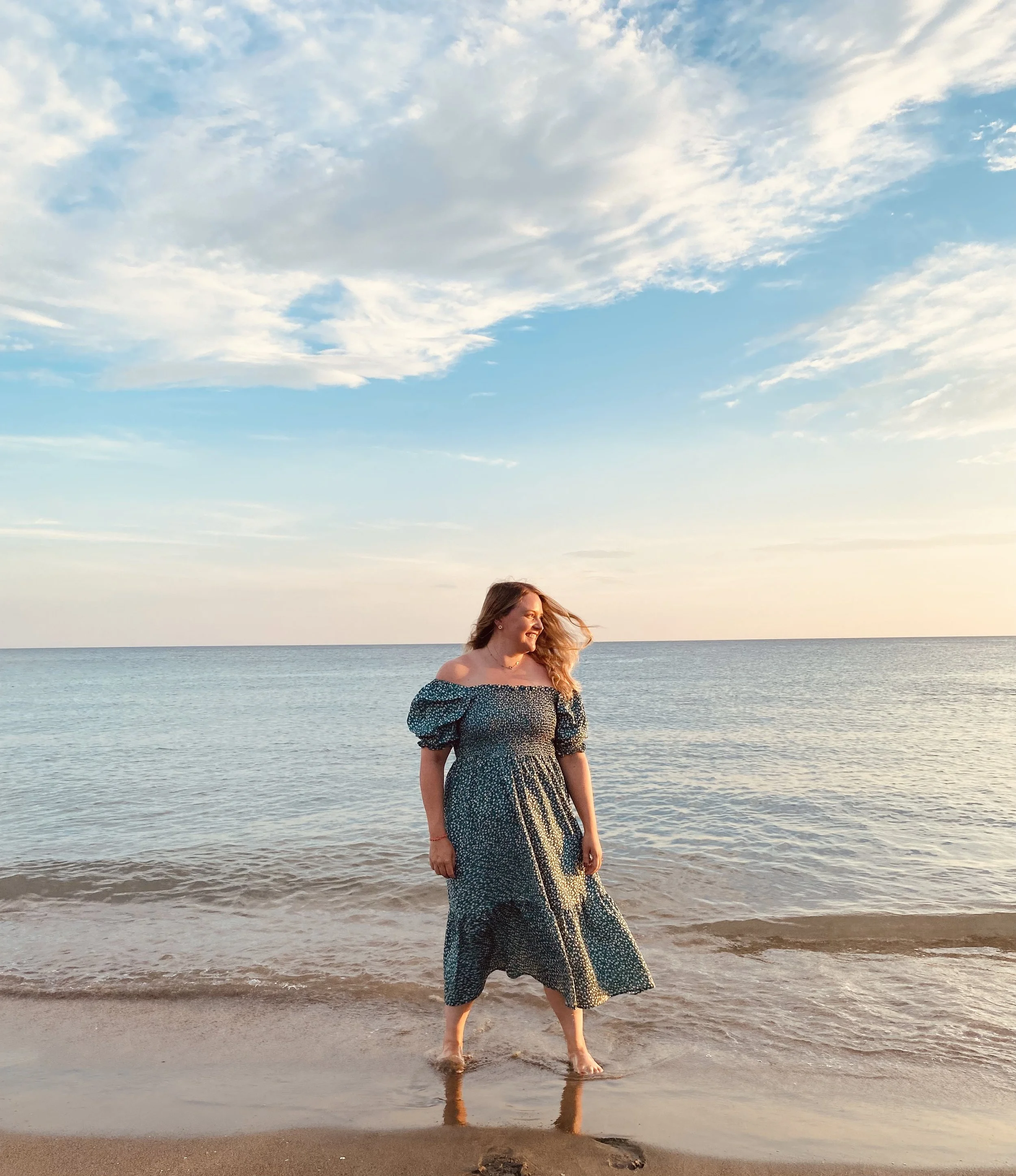 Eine lächelnde Frau in einem Sommerkleid steht barfuß im Wasser am Strand, mit Meer und Himmel im Hintergrund, während die Sonne untergeht.