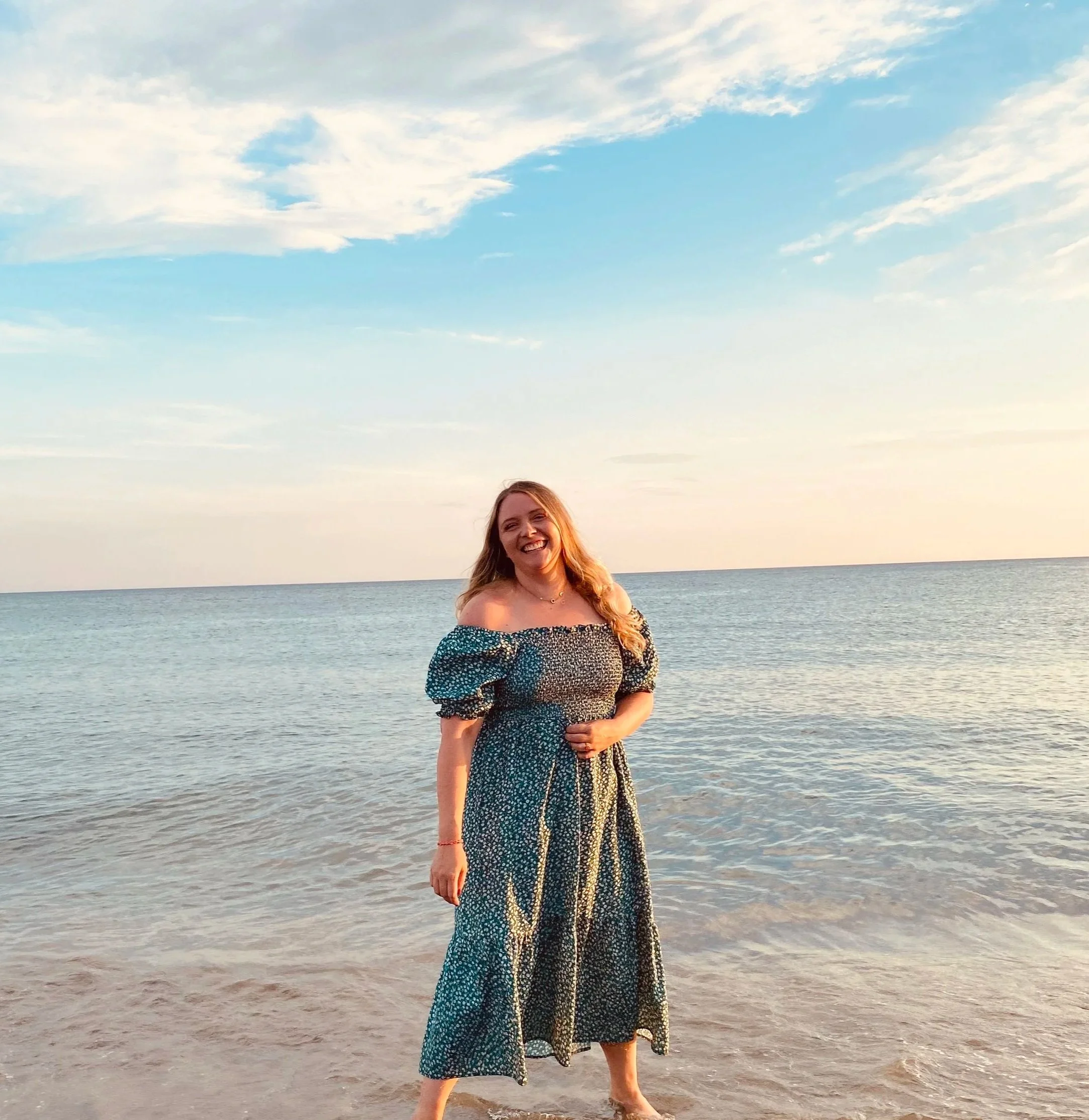 Eine junge Frau mit langen braunen Haaren trägt ein langes, schwarzes Outfit mit weißen Punkten am Strand bei Sonnenuntergang, mit Meer und Himmel im Hintergrund.