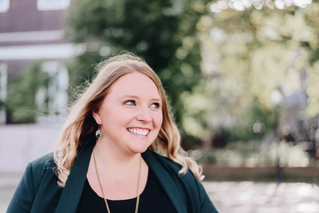 Lächelnde Frau mit blonde Haare, schwarzen Oberteil, schwarzen Blazer und Ohrringen, im Freien bei Sonnenschein, im Hintergrund Bäume und Gebäude verwischt.
