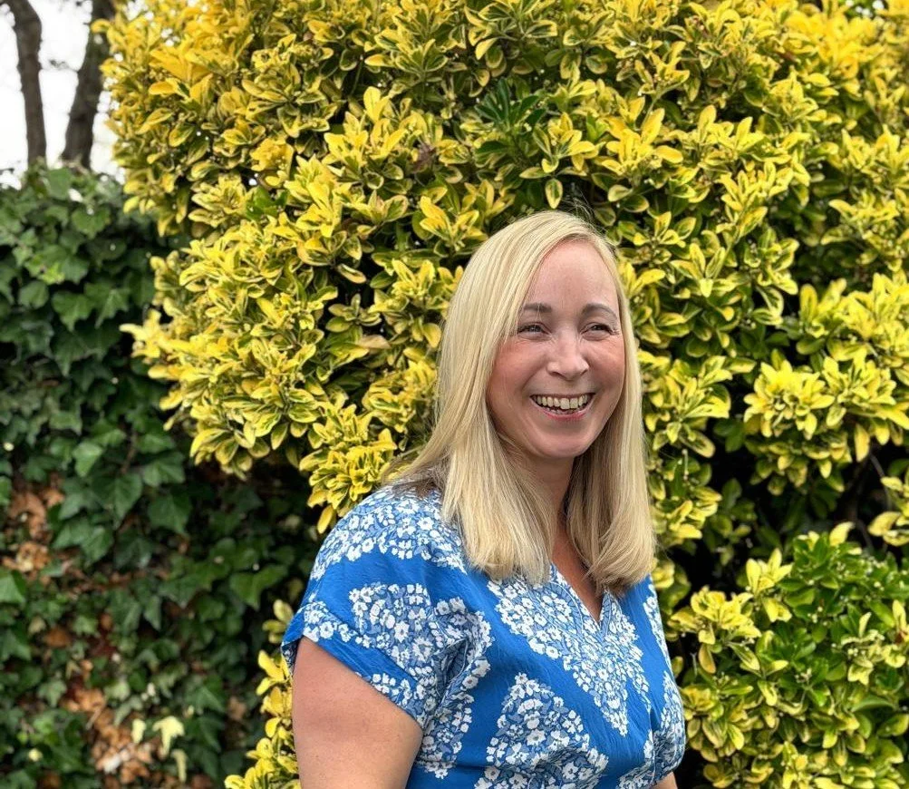 A smiling woman with blonde hair in a blue and white floral top standing in front of a yellow and green leafy bush.