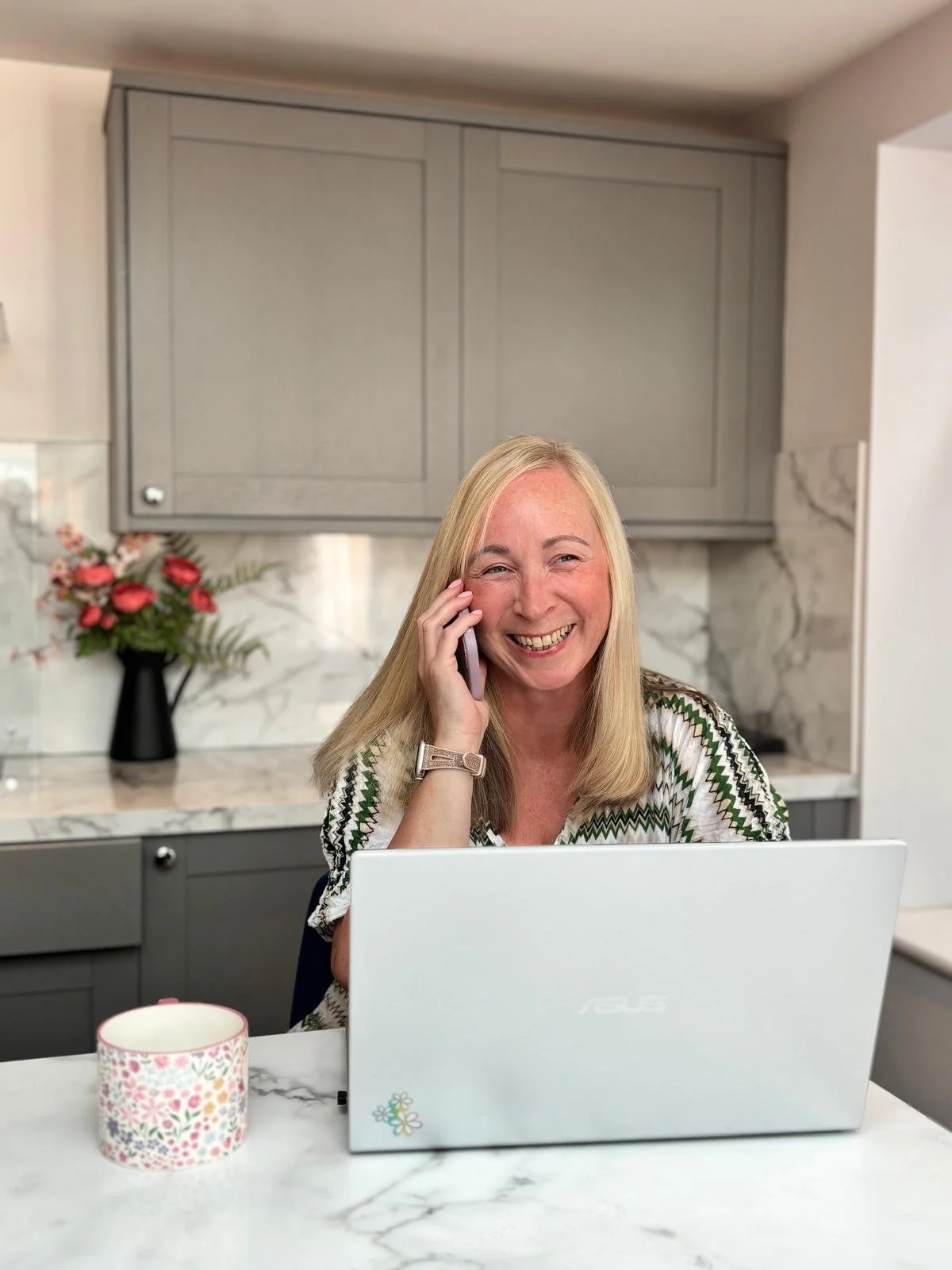 A woman with blonde hair smiling while talking on the phone at a kitchen counter, with a laptop in front of her and a vase of flowers in the background.