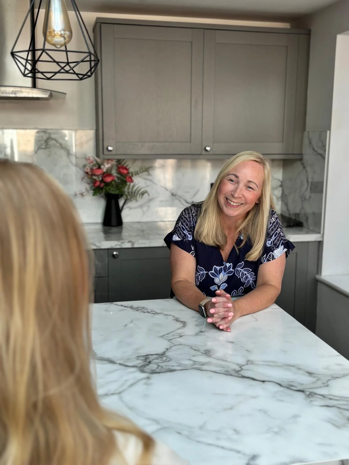 A woman with blonde hair smiling and sitting at a marble kitchen counter, with another person in the foreground. The kitchen has gray cabinets, marble backsplash, and a black vase with flowers in the background.