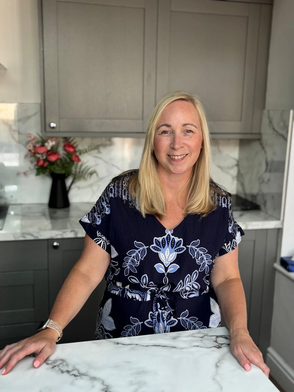 A woman with blonde hair smiling in a kitchen with gray cabinets and marble countertops, wearing a navy blue dress with white floral patterns.