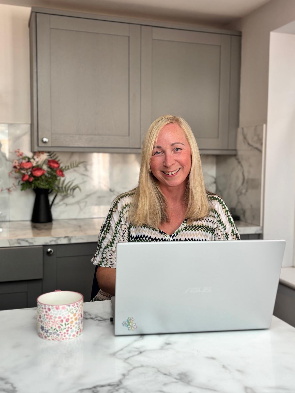 A woman with long blonde hair smiling while sitting at a kitchen counter with a laptop in front of her. There are pink flowers in a black vase and a mug on the counter. The background features gray cabinets and marble backsplash.