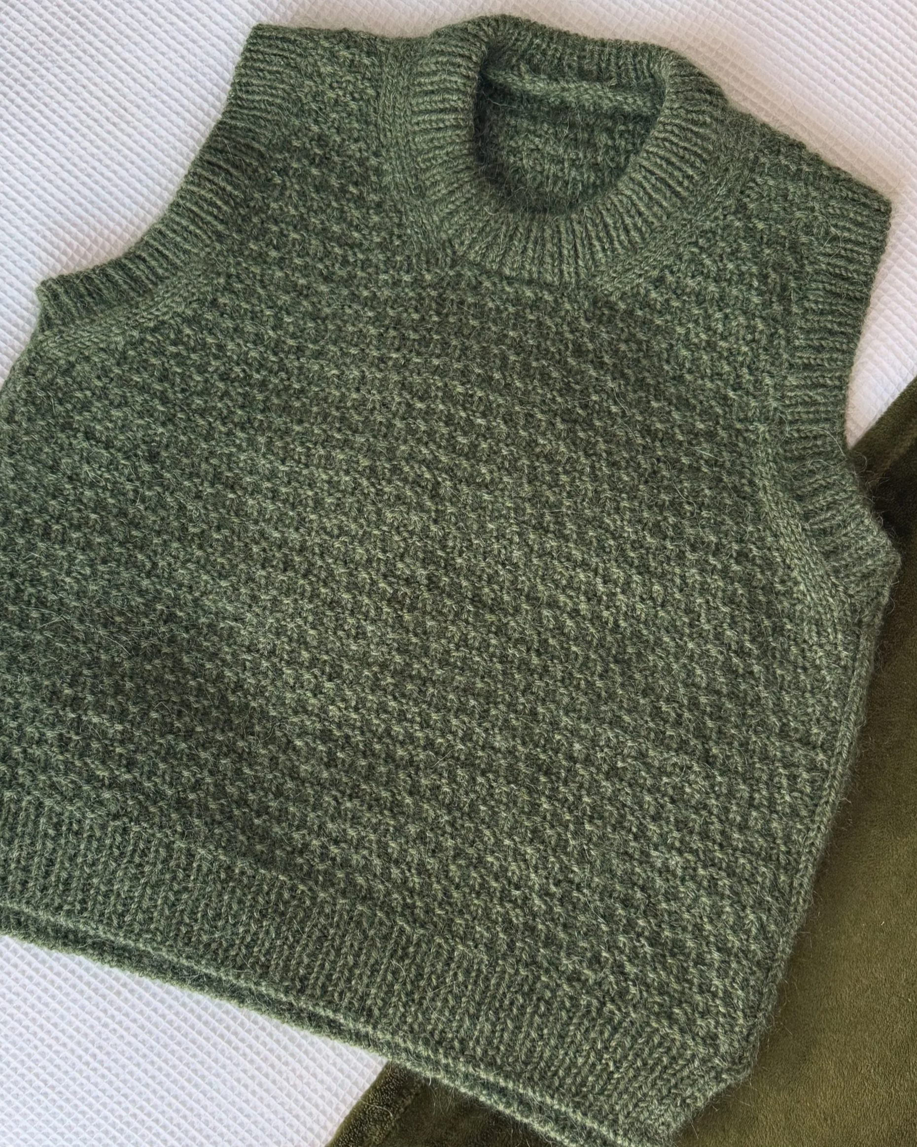 Tao Slipover, a knitting pattern by Hive Knits. Green knitted textured slipover or sweater-vest or vest with a ribbed crew neck in all-over Tao stitch texture laid out on a white textured surface.
