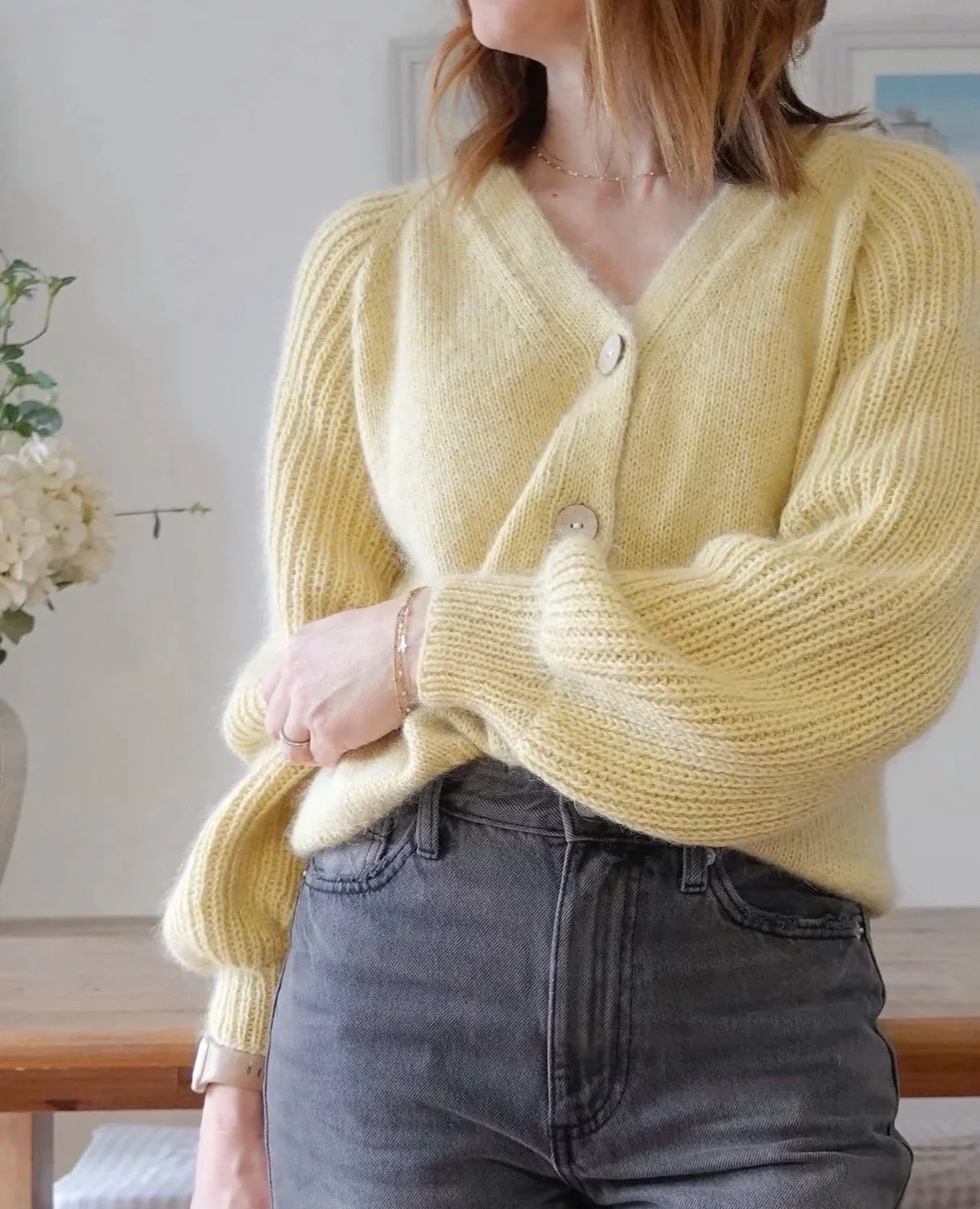 The First Cardigan, a knitting pattern by Hive Knits. A woman with shoulder-length red hair wearing a yellow v-neck knitted cardigan with large buttons, and statement textured Half Fisherman's Rib sleeves, and grey jeans,